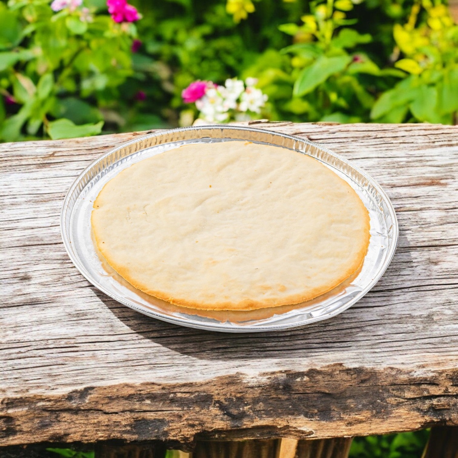A Rich's Pizza Crust Pan 10 Inch Gluten Free Par Baked crust rests on a silver tray atop a weathered wooden surface, framed by colorful flowers and greenery in the background.