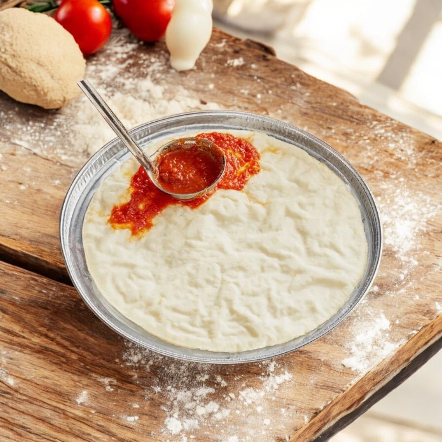 A metal bowl with pizza dough rests on wood as tomato sauce is ladled onto Rich's Pizza Crust Pan 10 Inch Gluten Free Par Baked. Nearby, fresh tomatoes, mozzarella, and a ball of flour-dusted dough wait their turn.