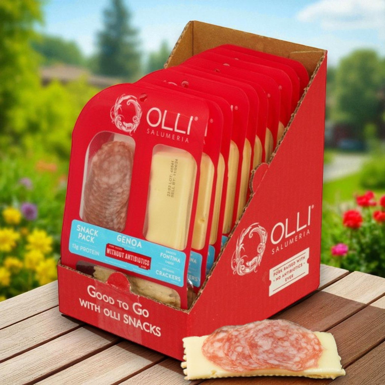A case of 10 Olli Salami Genoa Fontina Snack Packs (2 oz each) with crackers is displayed outdoors, featuring one opened high-protein snack pack in front.