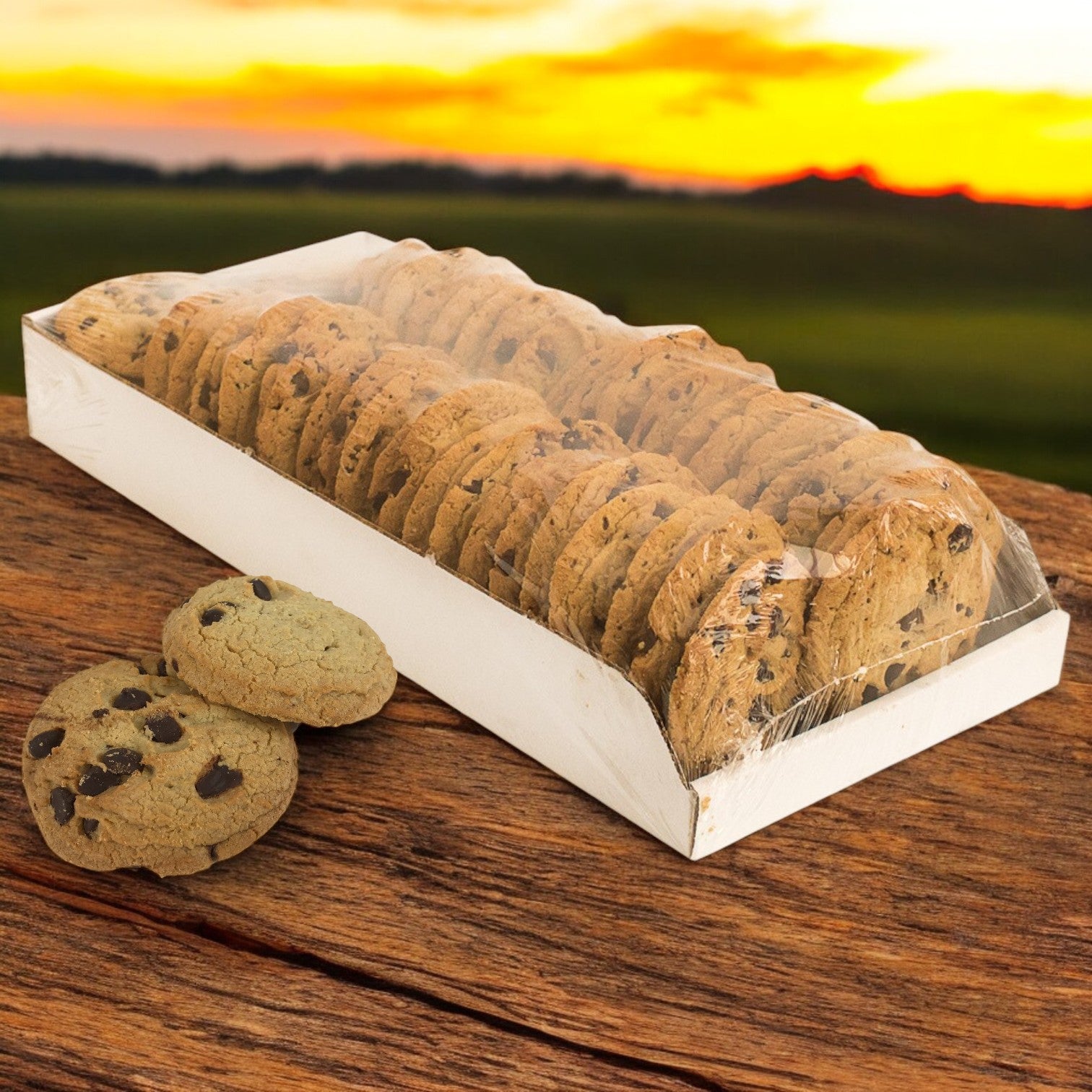 A long rectangular box of David's Cookies - Chocolate Chip Cookies (Baked, Thaw & Serve, 48/2 oz) sits on a wooden surface with a couple of treats beside it. A stunning sunset forms the backdrop, accentuating the inviting display.
