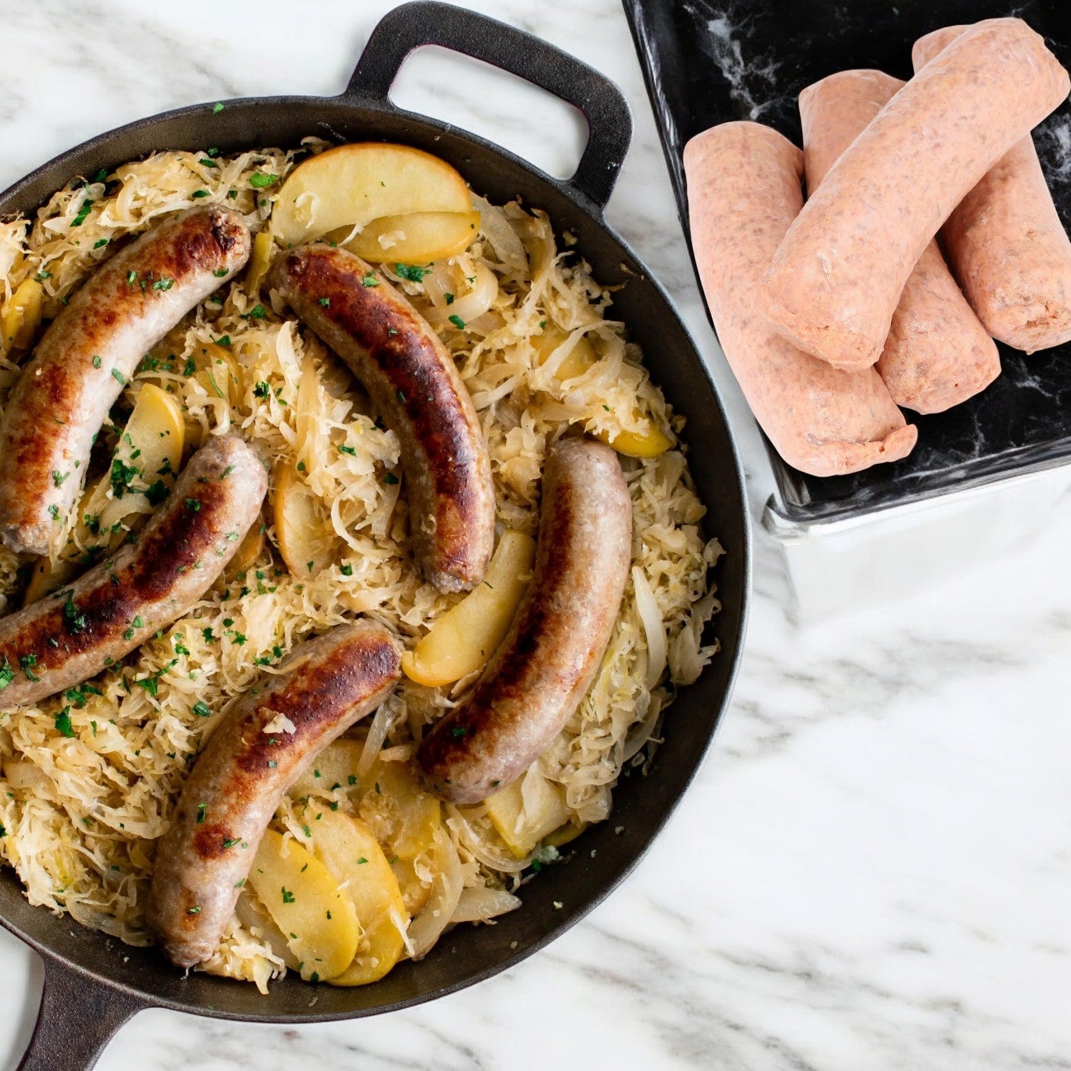 A skillet with browned Beyond Meat Sausage Vegetarian Original Brat Links, sliced potatoes, and sauerkraut sits alongside four uncooked bratwursts on a plate.