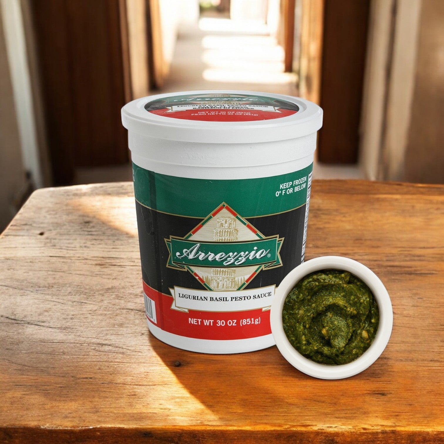 On a wooden table sits a package from Sysco: Arrezzio Imperial Ligurian Basil Pesto Sauce, offering an authentic Italian taste. One of the three 30 oz containers is open beside a small bowl filled with the vibrant green pesto.