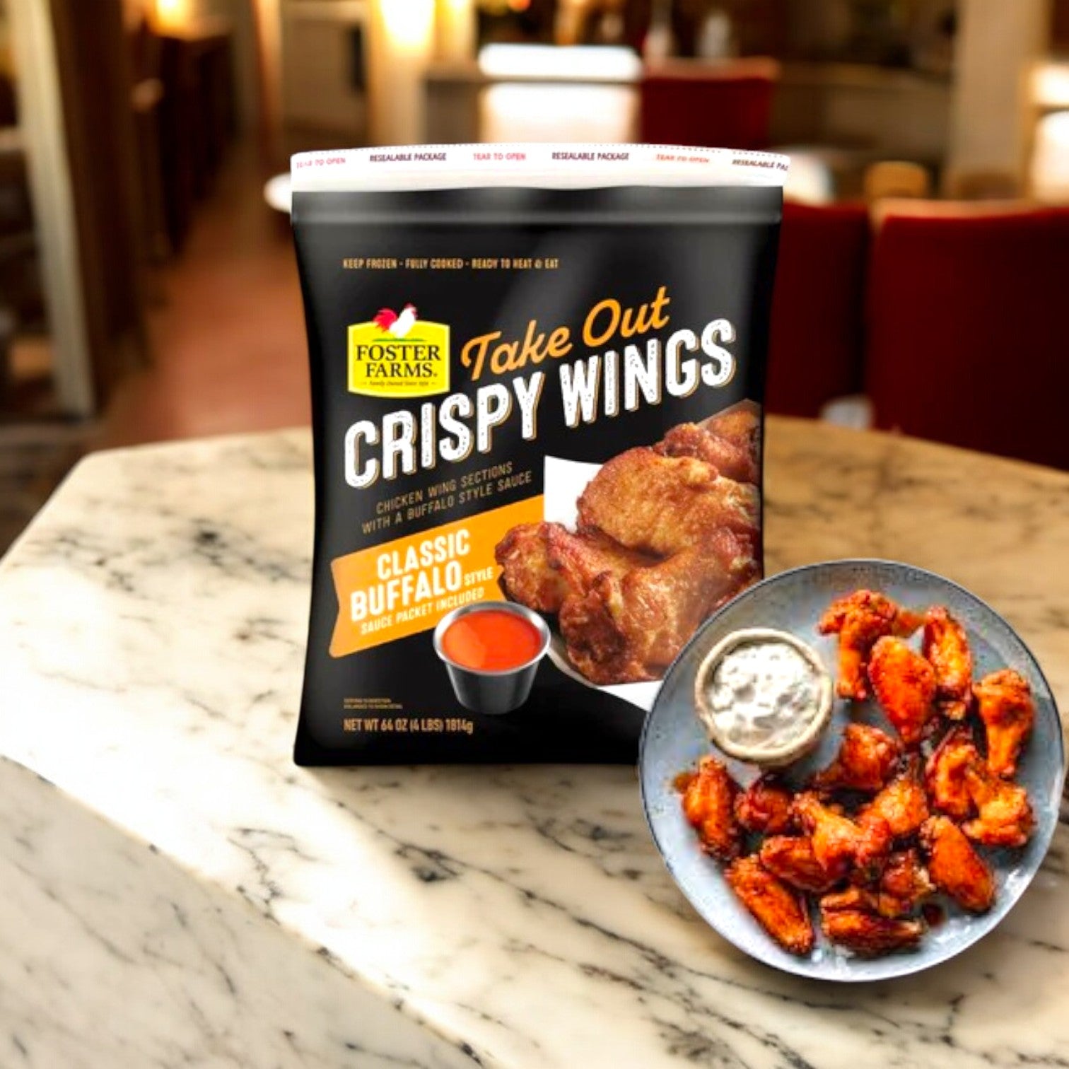 A package of Foster Farms Take Out Crispy Chicken Wings in Classic Buffalo, 4 lbs, sits on a marble surface, next to a plate of wings and a small dipping sauce cup.