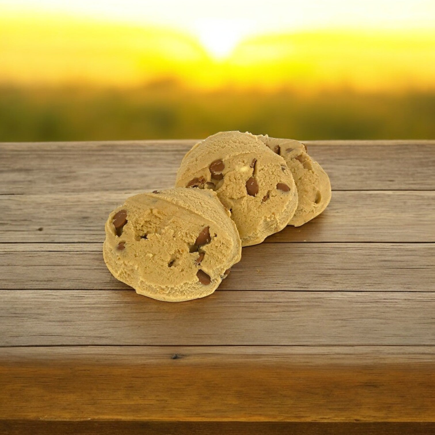 Three peanut butter cookies from David's Cookies Dough rest on a wooden surface, with a blurred sunset providing the perfect backdrop.