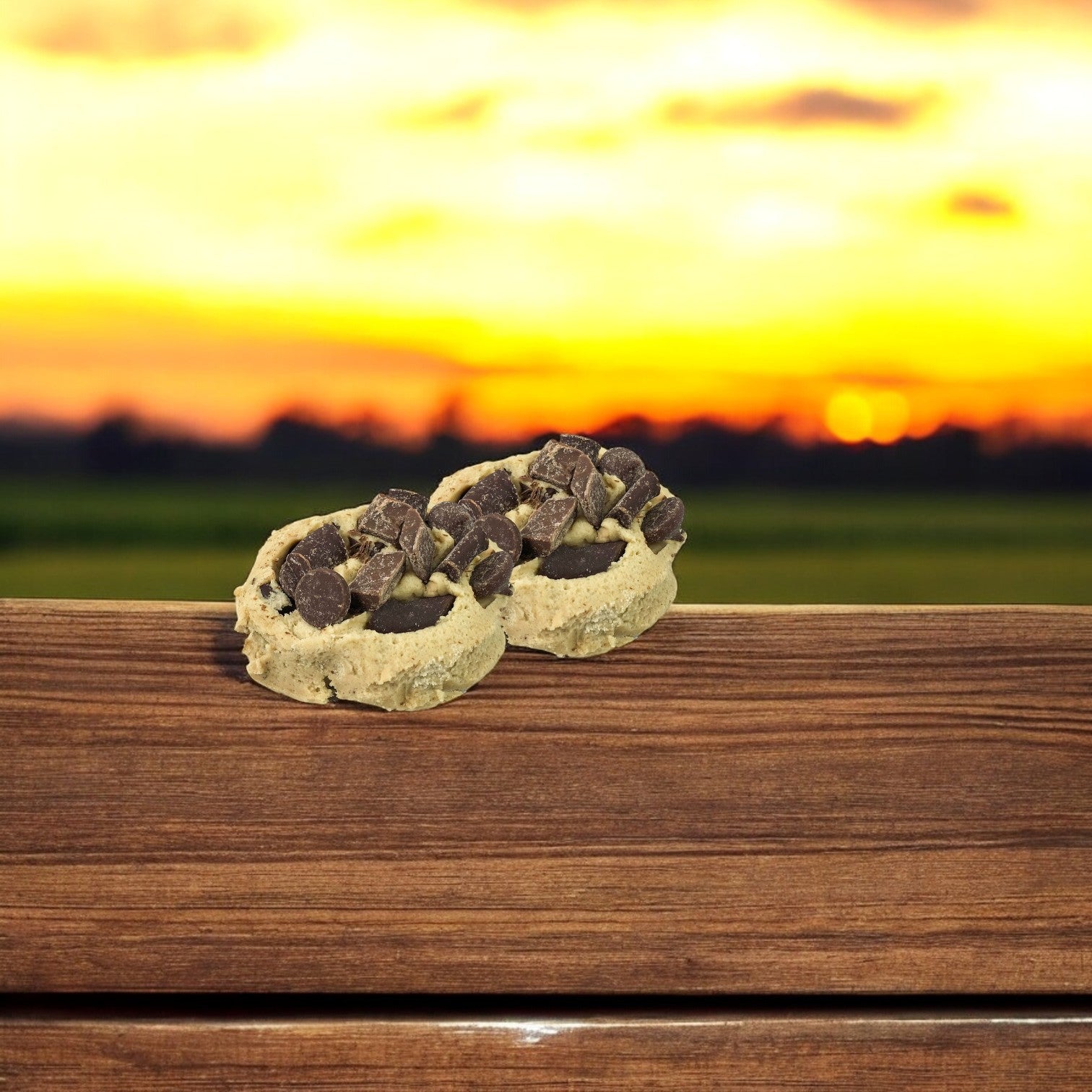 Two of David's Cookies' Triple Chocolate Dough cookies, brimming with extra chocolate pieces, lie on a wooden surface against a sunset backdrop—a perfect treat for chocolate lovers.