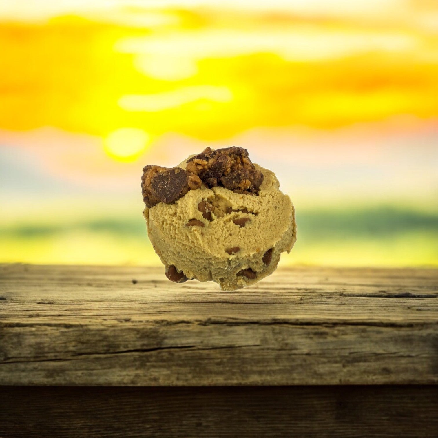 A scoop of pistachio ice cream paired with David's Cookies Dough Cookie - Reese's Peanut Butter Chunk rests on a wooden surface, set against a vibrant sunset backdrop.