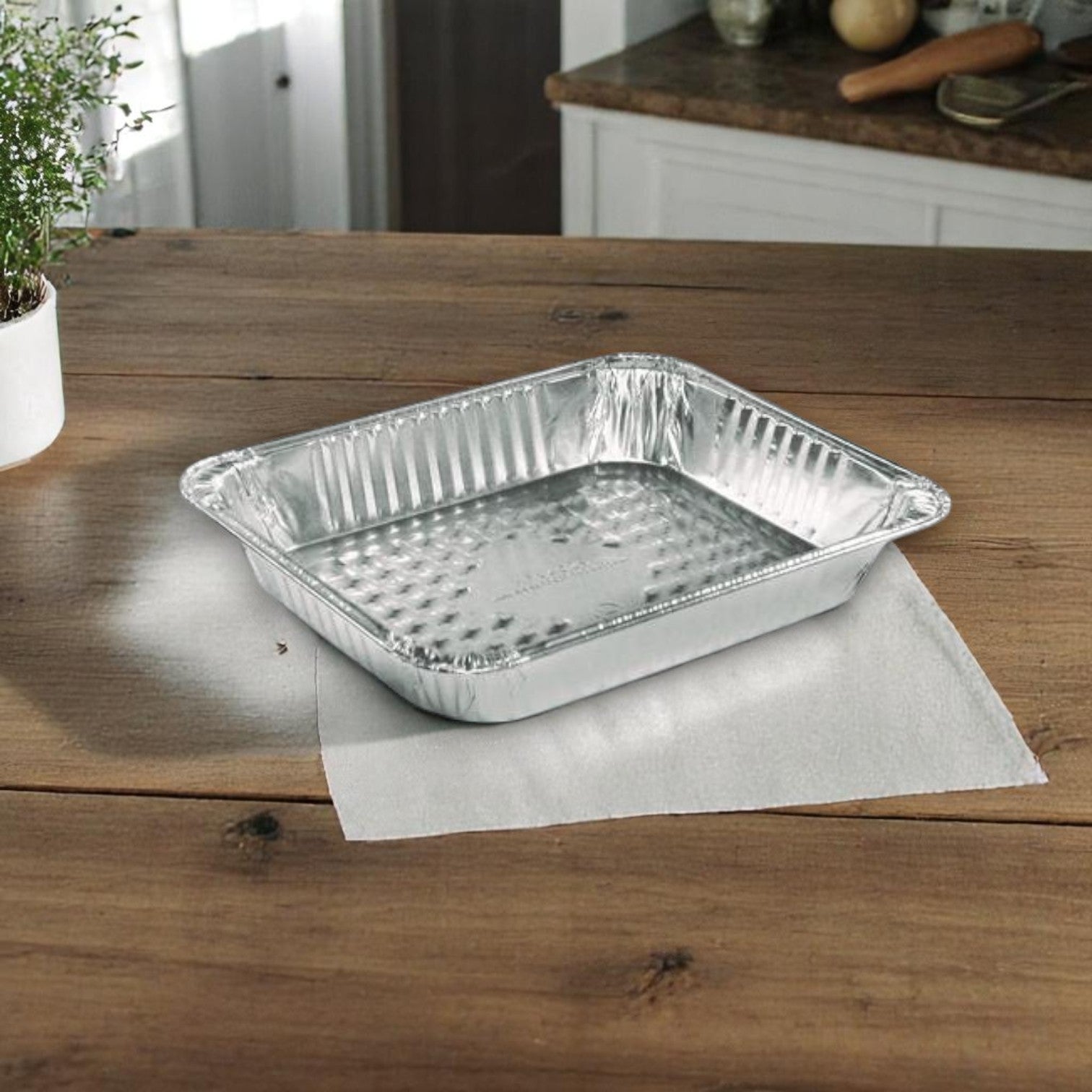 A Handi-Foil of America HFA Half-Size Deep Steam Table Pan from the 12 Pack sits on a paper towel on a wooden kitchen table, with a plant and kitchen items in the background.