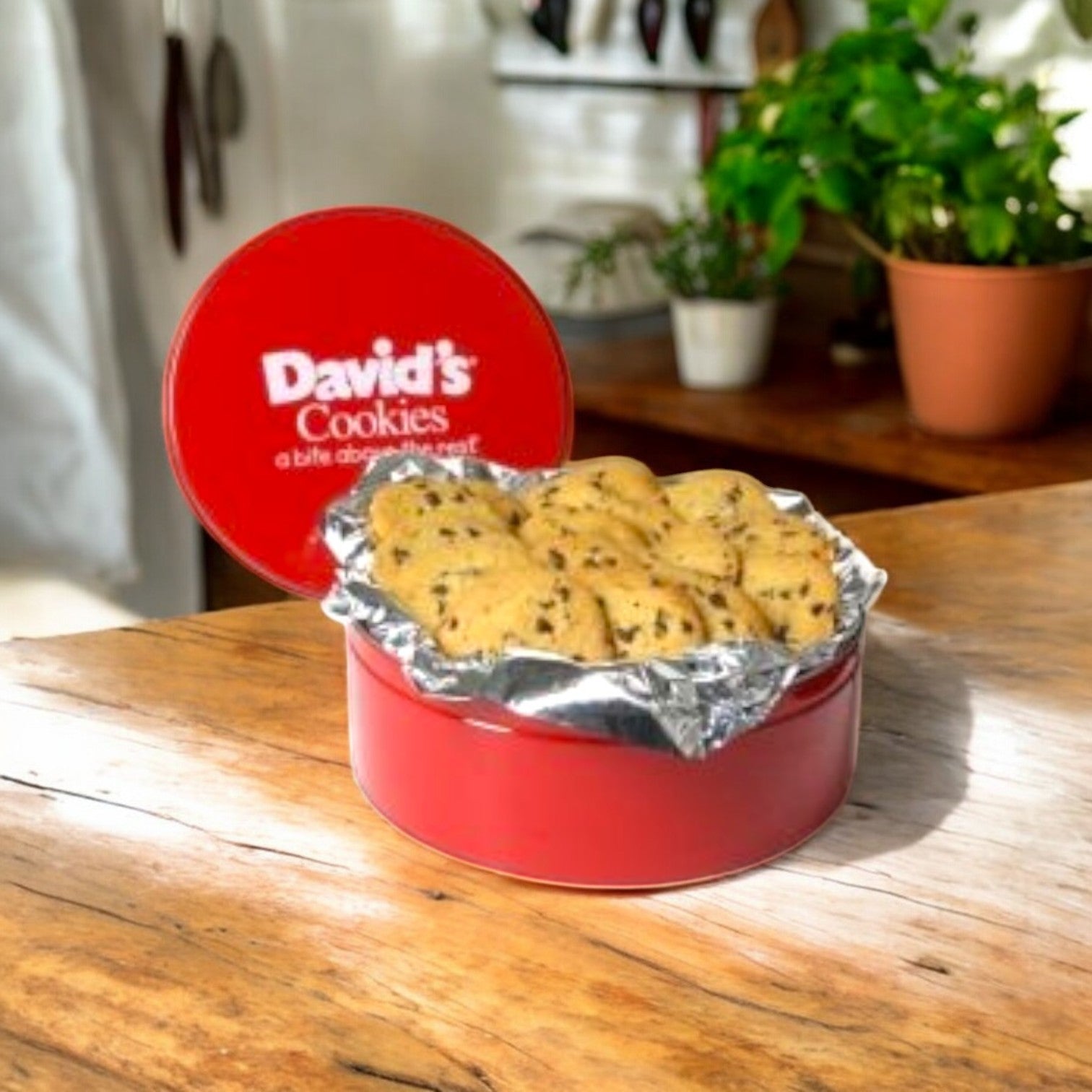 On a wooden table, open the red tin labeled "David's Cookies," revealing David's Cookies - Chocolate Chip Cookie Dough (1.5 oz., 120 Count) on foil. A potted plant and kitchen utensils in the background highlight the charm of fresh cookies ready for baking.
