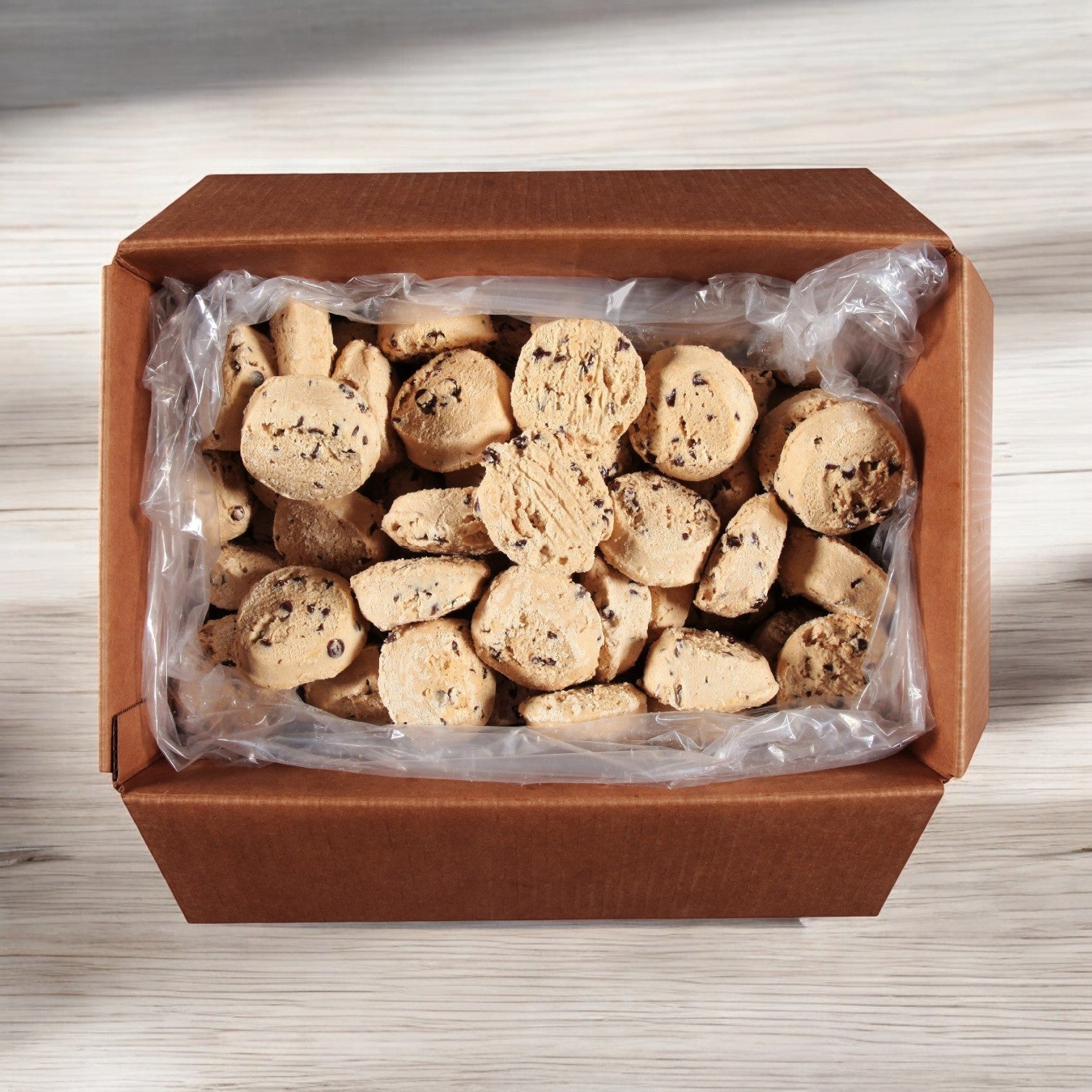 A bulk pack of Otis Spunkmeyer Dough Cookie Chocolate Chip 80/4 oz. cookies is nestled in an open cardboard box with a plastic lining, each cookie gleaming temptingly in its random arrangement.