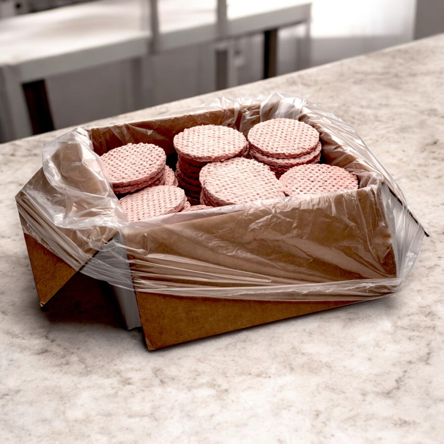 A cardboard box lined with plastic contains stacks of round, pink wafer cookies, reminiscent of the precision used in creating a Fire River Farms Imperial by Sysco Halal Beef Patty – 80 Pack (4 oz Each). The box rests elegantly on a marble countertop.