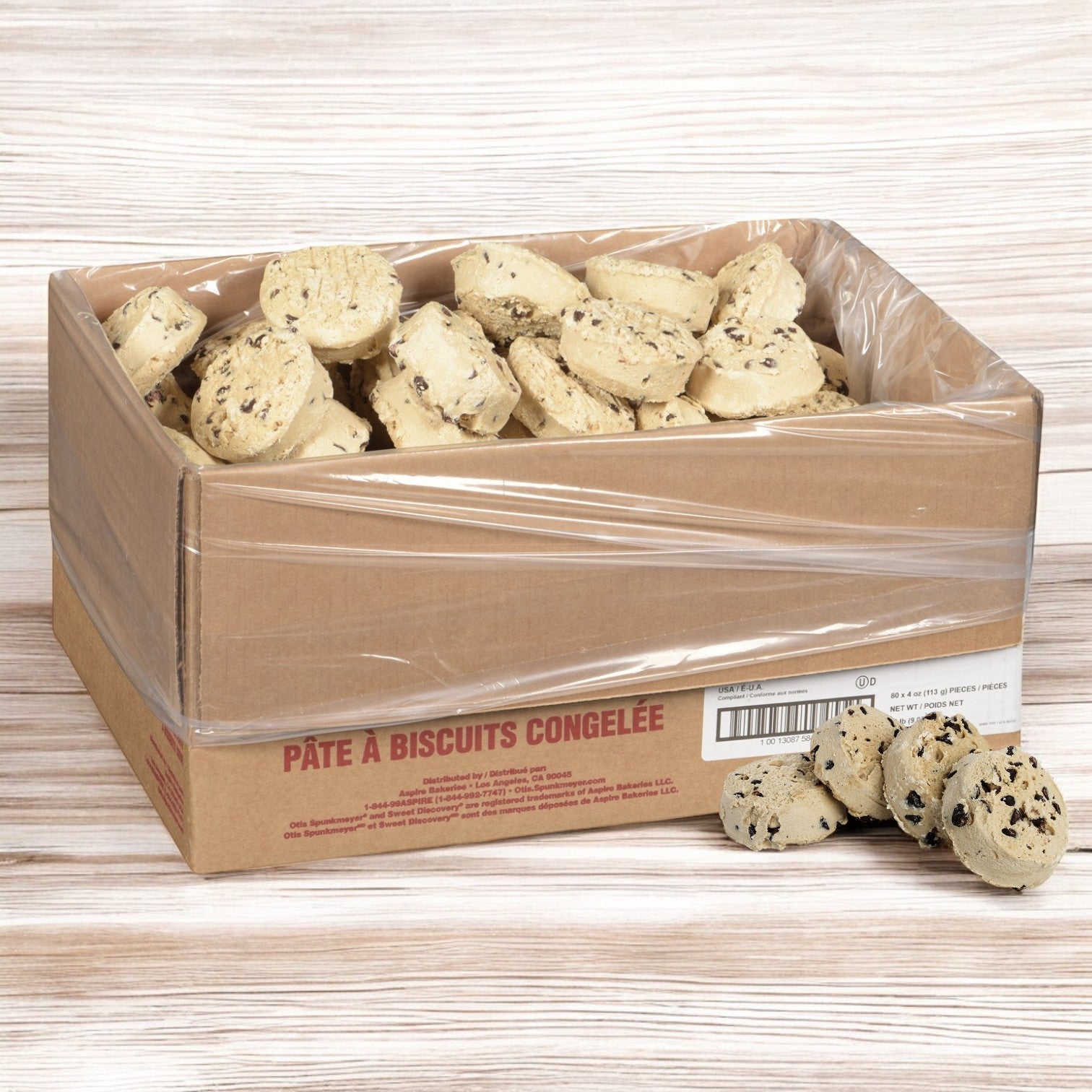 An Otis Spunkmeyer bulk pack of Chocolate Chip Cookie Dough, containing 80 pre-portioned 4 oz. frozen pieces, sits in a cardboard box with some dough ready on a wooden surface for baking.