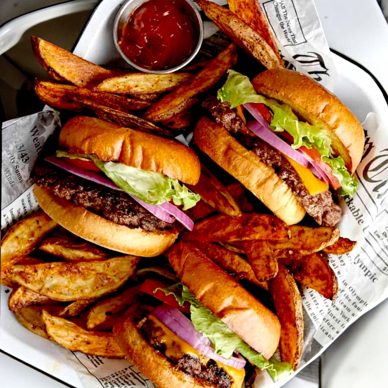Three burgers with Fire River Farms Imperial Halal beef patties, lettuce, tomato, onion, and cheese are served with seasoned potato wedges and ketchup on a newspaper-lined plate.