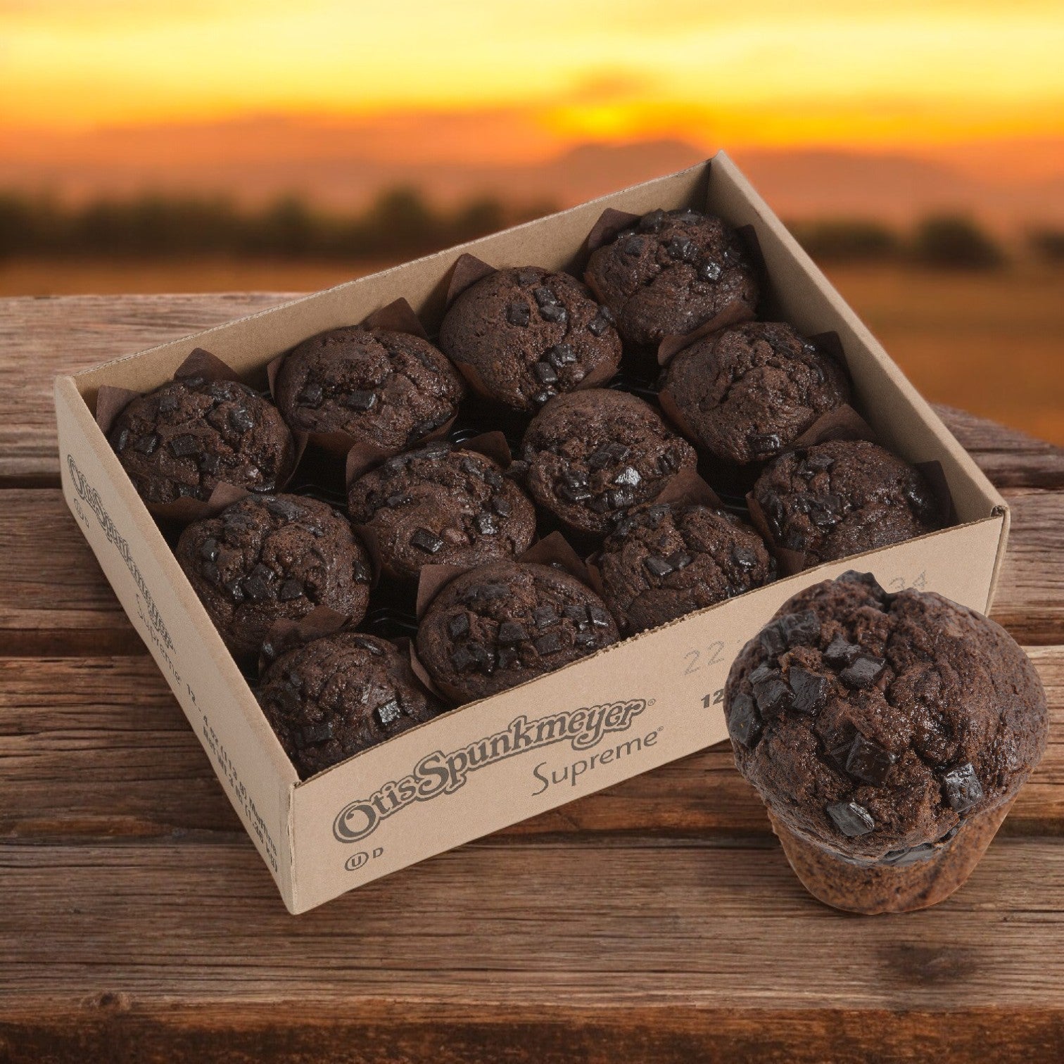 A bulk pack of Otis Spunkmeyer Muffin Chocolate Chunk Supreme 24/4 oz. sits on a wooden table, with individually wrapped muffins for freshness. One muffin is outside the box against a beautifully blurred sunset backdrop.
