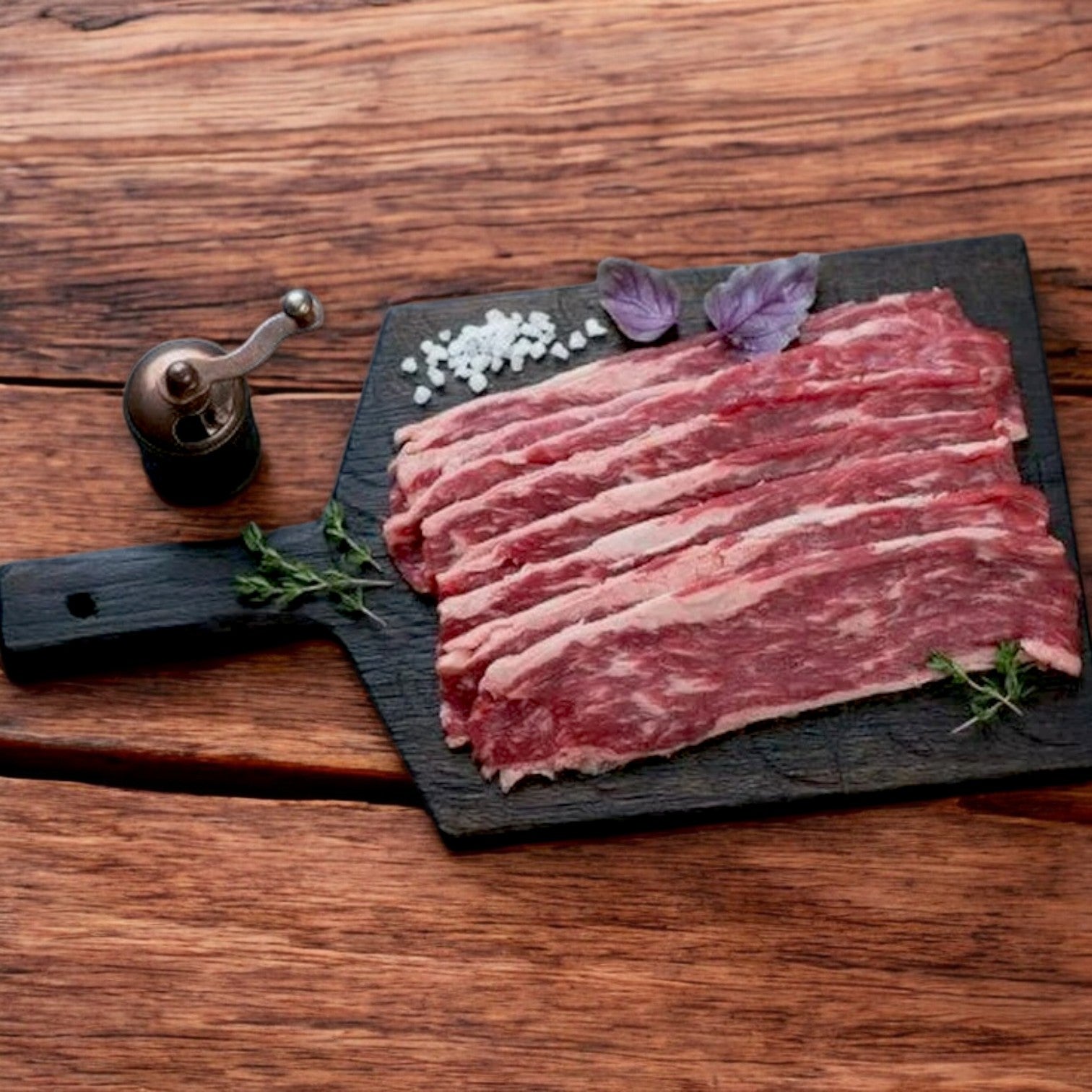 Greco & Sons Halal Beef Bacon Strips on a dark cutting board, garnished with herbs. Salt and a pepper grinder are nearby.