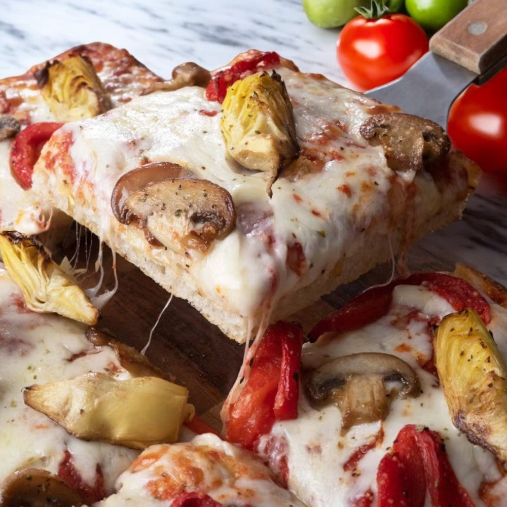 A close-up of a Massa Pizza Co. Roasted Veggie Sicilian Bakery Pizza slice, featuring melted cheese, mushrooms, artichokes, and tomatoes on a wooden board. Whole tomatoes and a pizza cutter sit in the background, emphasizing the artisanal dough that perfects this roasted veggie delight by Massa Pizza.