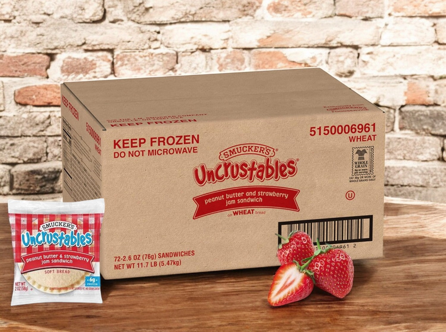 A box of Uncrustables 72 count - Strawberry Jam by Uncrustable Smuckers sits on a wooden surface, surrounded by fresh strawberries and a packaged sandwich filled with sweet strawberry jam and creamy peanut butter.