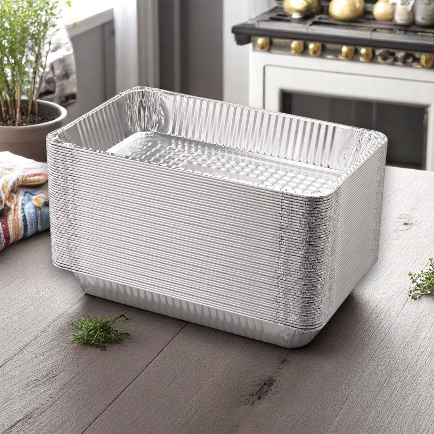 A stack of Handi-Foil of America HFA Deep Full-Size Steam Table Pans (10-count pack) rests on a wooden kitchen table near herbs and a stove in the background.