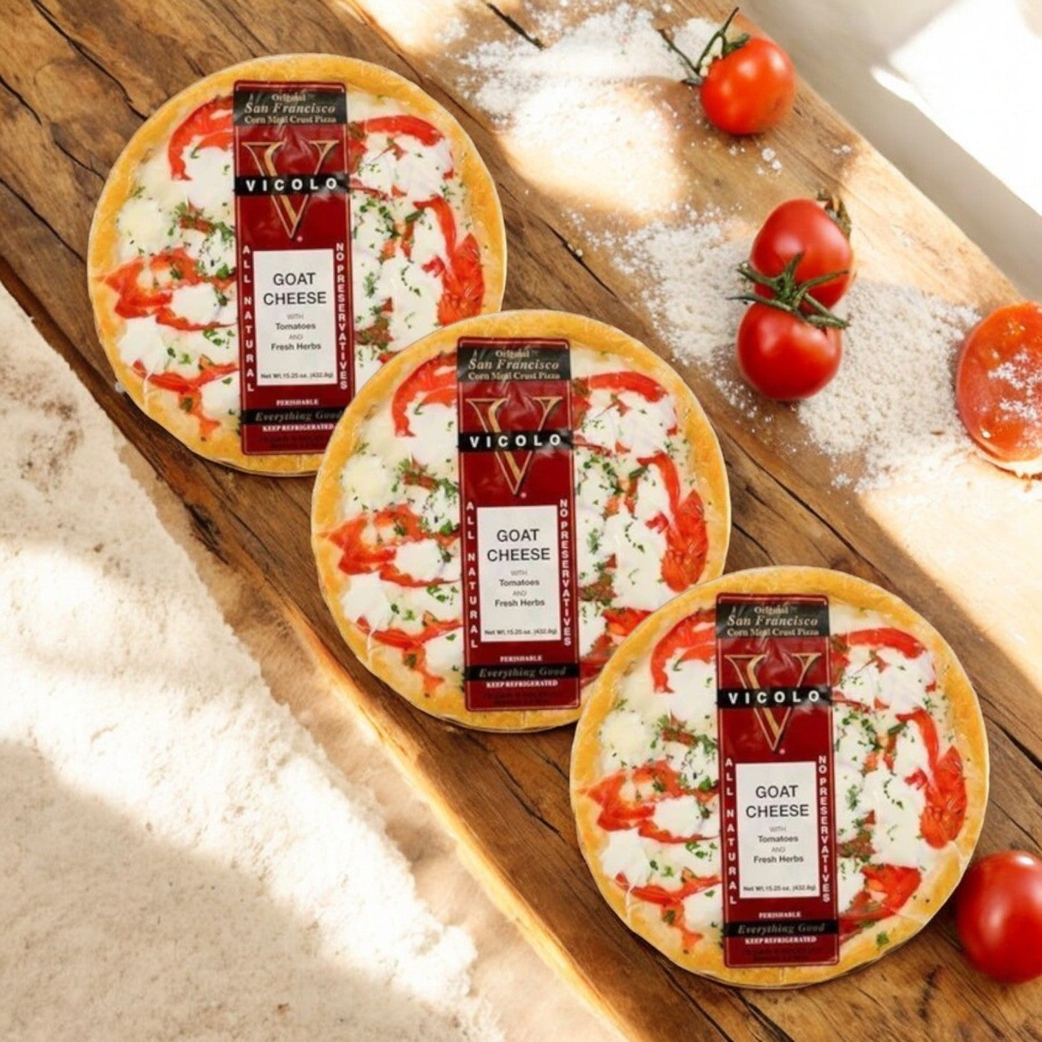 Three Vicolo Goat Cheese Pizzas are artistically placed on a wooden surface, surrounded by flour and fresh tomatoes. The artisanal crust promises crisp perfection with every bite.