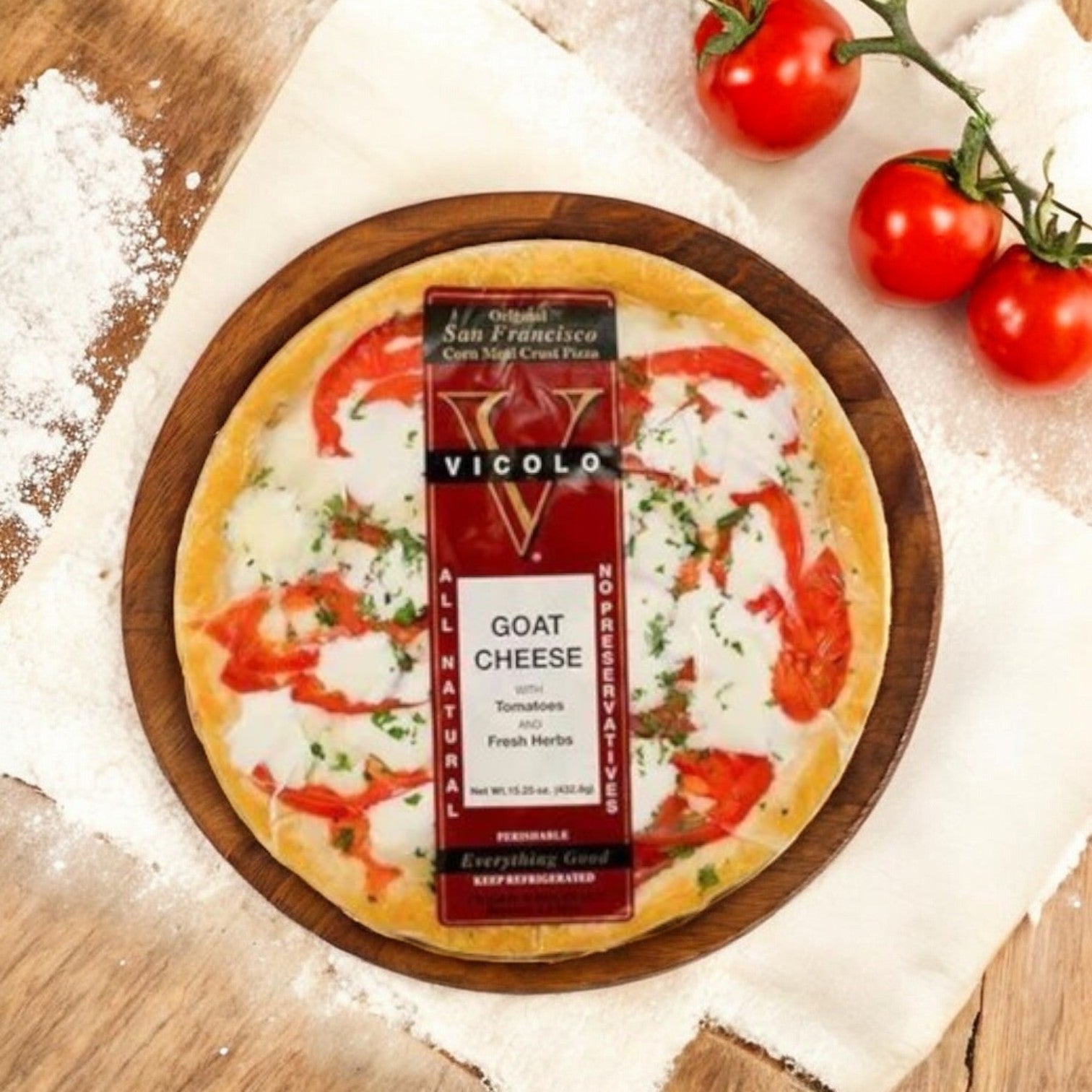 A Vicolo Goat Cheese Pizza-1 Count, adorned with tomatoes and herbs, sits on a wooden board highlighting its artisanal crust. Surrounded by flour and tomatoes on rustic wood, it epitomizes gourmet pizza delight.