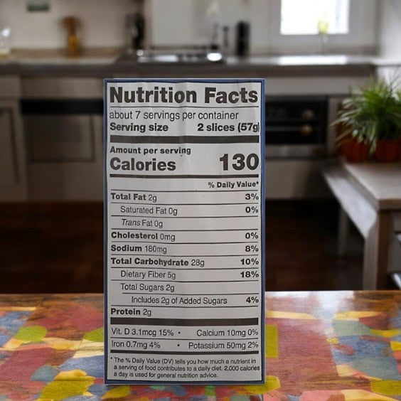 The Schar Bread Artisan Baker Gluten Free White has a nutrition label ideal for those with dietary restrictions, featuring 130 calories per serving, 2g total fat, 180mg sodium, and 2g total sugars—perfect beside your artisan baker's setup in a blurred kitchen background.