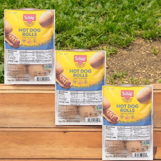 On a wooden table outdoors are three packs of Schar Rolls Hot Dog Gluten Free, each containing 4 rolls and weighing 8 oz (228g). Ideal for those with gluten sensitivities or celiac disease, they offer the classic taste worry-free.