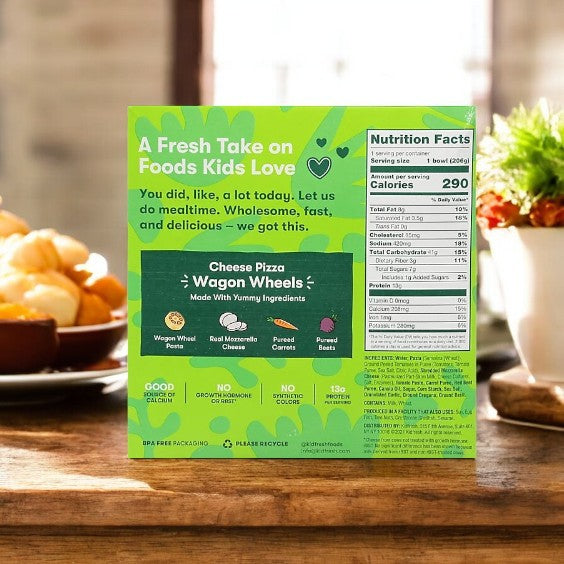 The Kidfresh Wagon Wheels Cheese Pizza, 7.25 oz., highlights nutrition facts and ingredients, emphasizing its organic whole wheat crust. The sunny scene with a window and oranges suggests hidden veggies in this wholesome pizza treat.