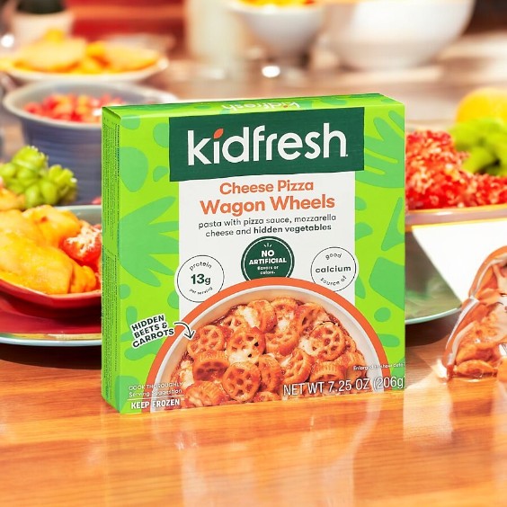 The Kidfresh Wagon Wheels Cheese Pizza, 7.25 oz., showcases pasta on its box and features 13g of protein, no artificial flavors, 9g of calcium, hidden vegetables like herbs and carrots, and an organic whole wheat crust. Packaging is displayed on a table with food in the background.
