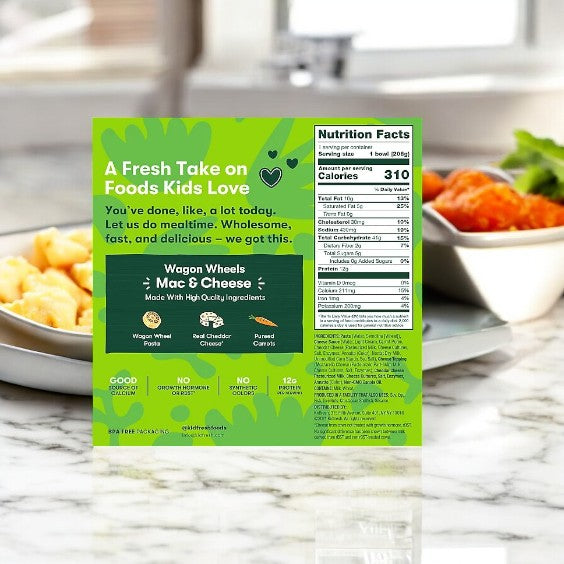 A box of Kidfresh Wagon Wheels Mac & Cheese, 7.25 oz.- 1 Count, is on the counter, highlighting its organic pasta and hidden veggies with clear nutritional details.