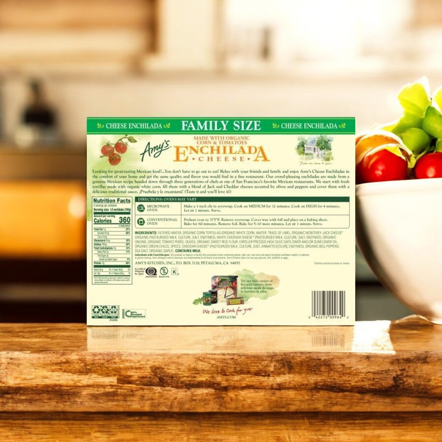 Back of a family-size box of Amy's Kitchen Cheese Enchilada, 9 oz., with organic corn tortillas on a kitchen counter, set against a backdrop of a bowl of tomatoes.