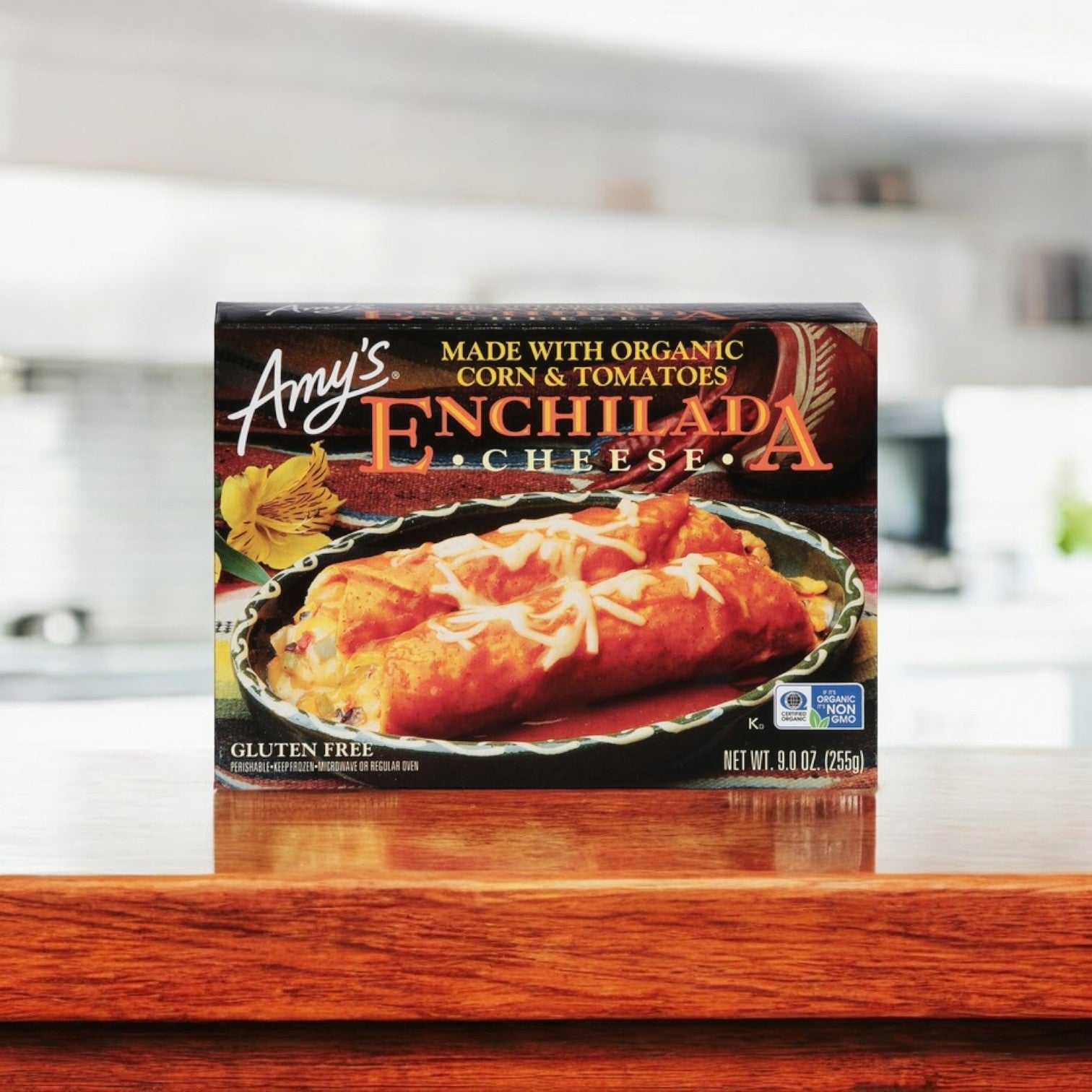 On a wooden countertop sits a box of Amy's Kitchen Cheese Enchilada, 9 oz., marked as vegetarian-friendly, made with organic corn and tomatoes, gluten-free, and non-GMO.