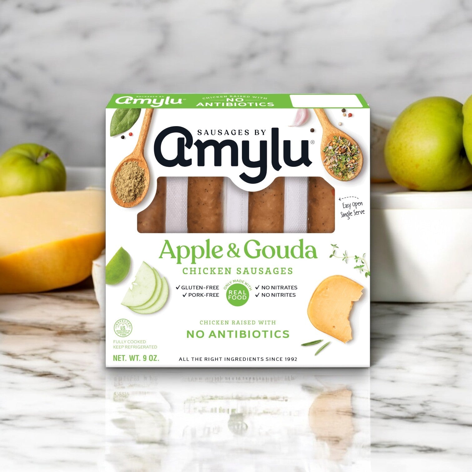 On a marble surface sits the box of Amylu Apple and Gouda Chicken Sausage - 3 Count, complete with cheese, an apple, and a garlic clove. This fully cooked sausage is proudly gluten-free with no antibiotics.