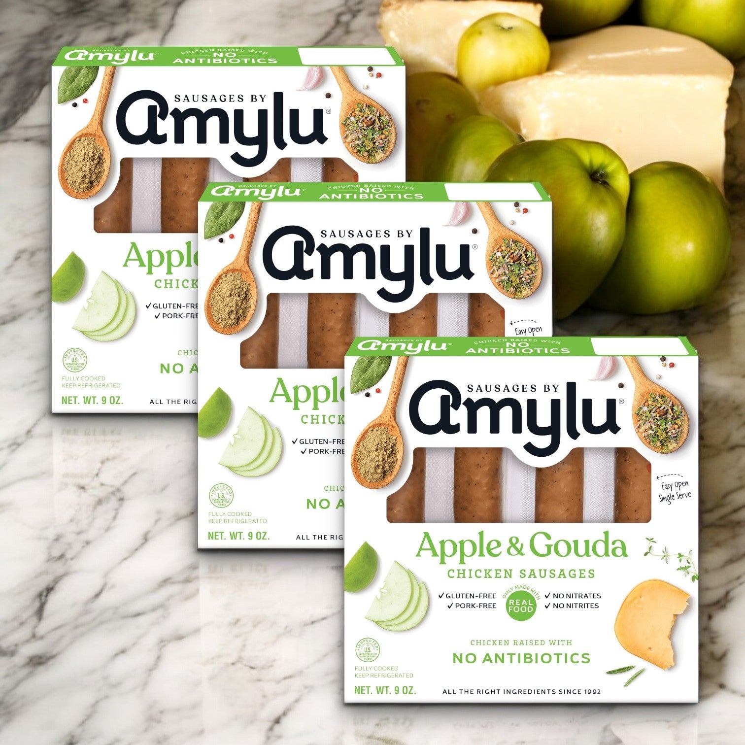 Three packs of Amylu Apple and Gouda Chicken Sausage - 3 Count are on a marble surface with green apples and cheese perfectly displayed behind them. Enjoy this fully cooked gourmet sausage delight from Amylu.
