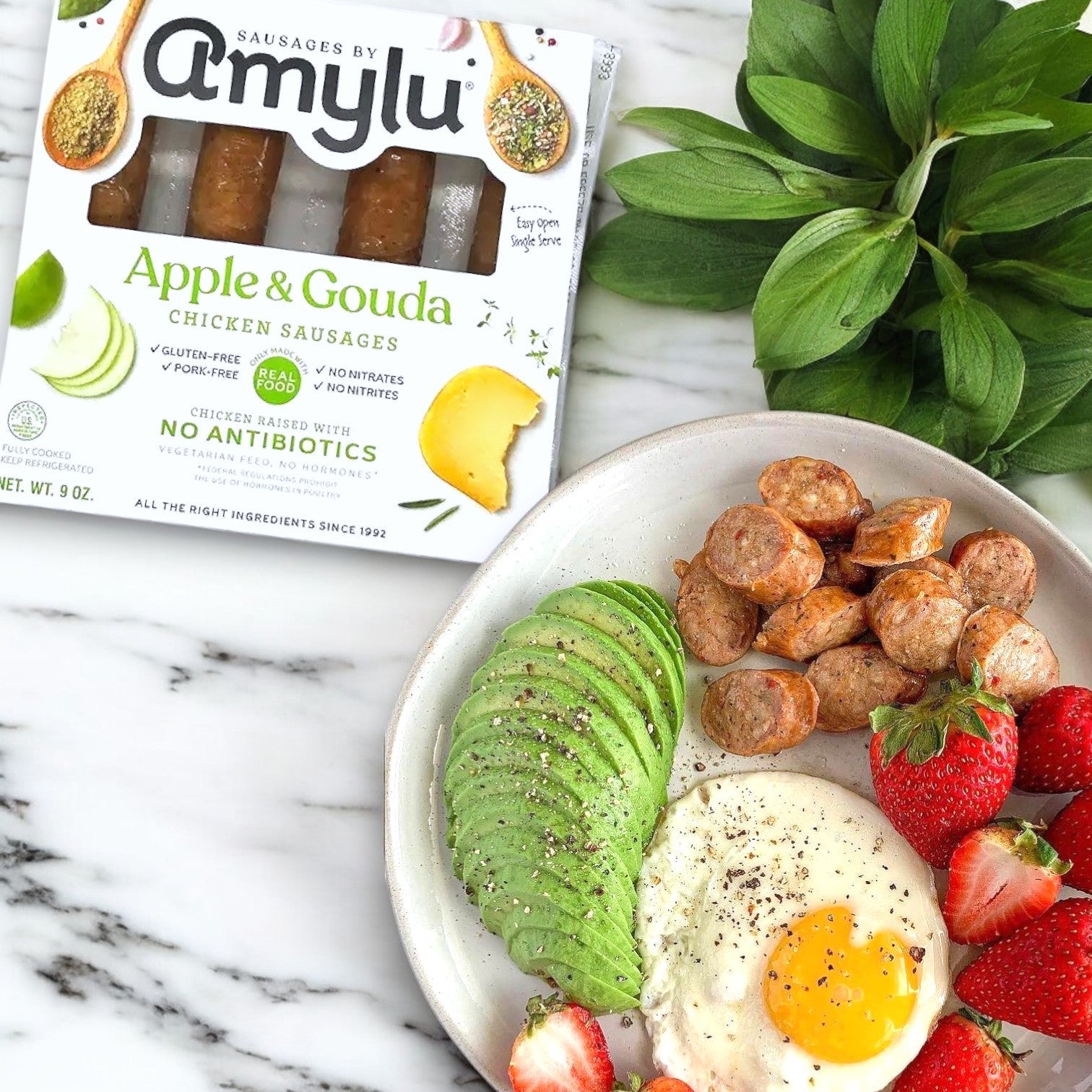Amylu Apple and Gouda Chicken Sausage - 1 Case adds a gourmet twist next to sliced avocado, sausages, a fried egg, and strawberries on a marble surface.