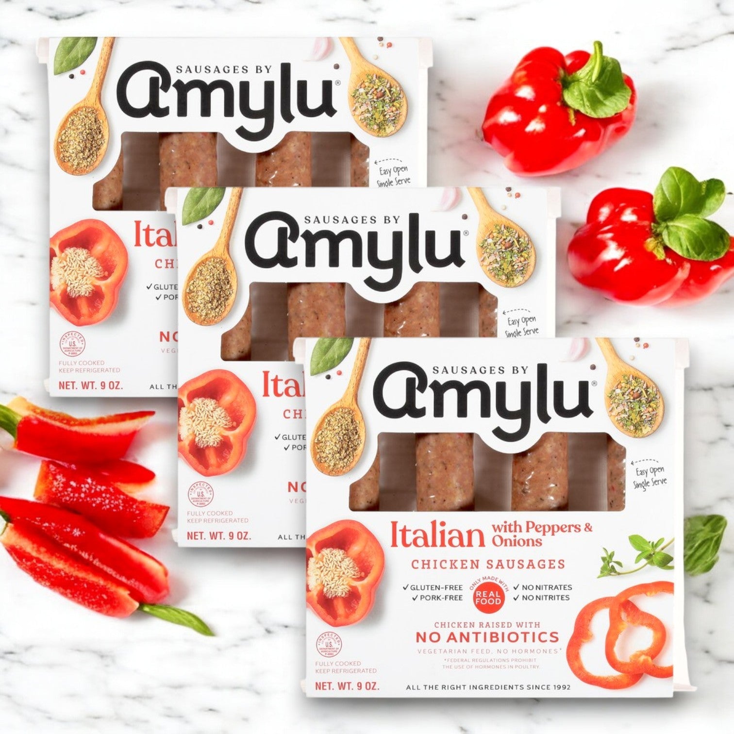 Three packs of Amylu Italian Chicken Sausage with Peppers & Onions are elegantly displayed on a marble surface, surrounded by sliced red peppers and fresh basil leaves.