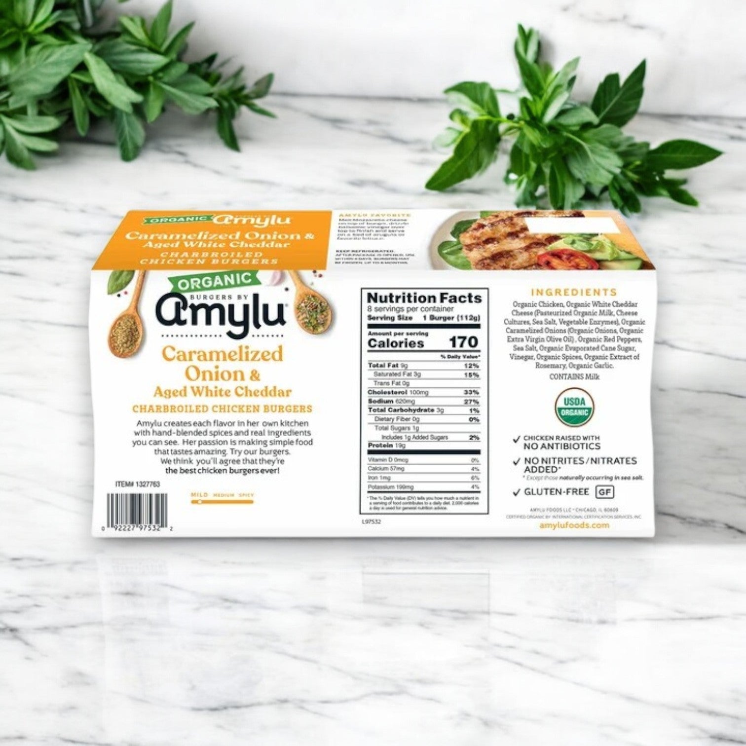 The front view of the 32 oz, 24-count Amylu Organic Chicken Burger with Caramelized Onion & Aged White Cheddar box highlights nutritional facts and ingredients alongside the prominent USDA organic label, set against a backdrop of scattered green herbs.