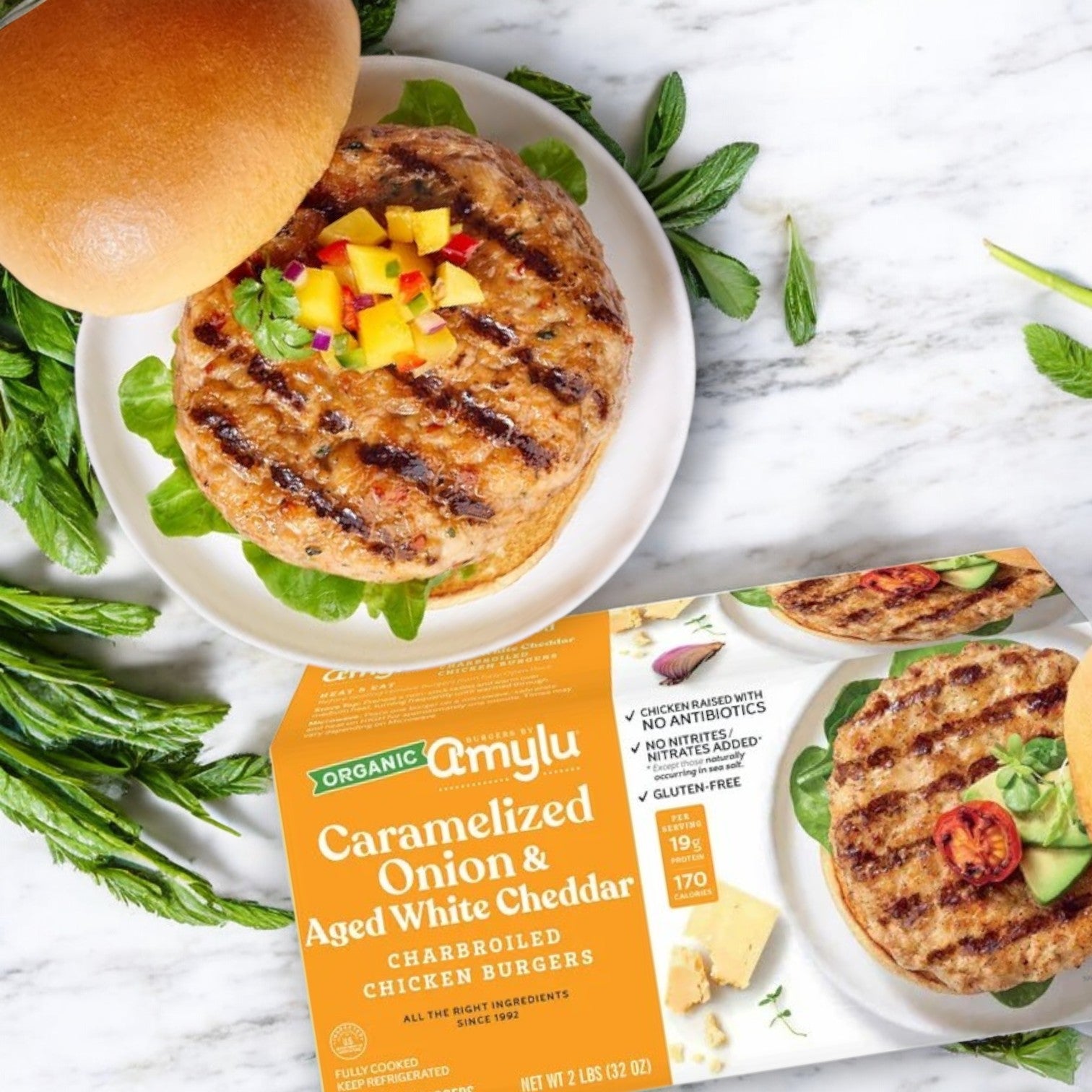 A chicken burger topped with mango salsa is on a plate next to an "Amylu Organic Chicken Burger with Caramelized Onion & Aged White Cheddar" package, surrounded by fresh greens and visible branding.