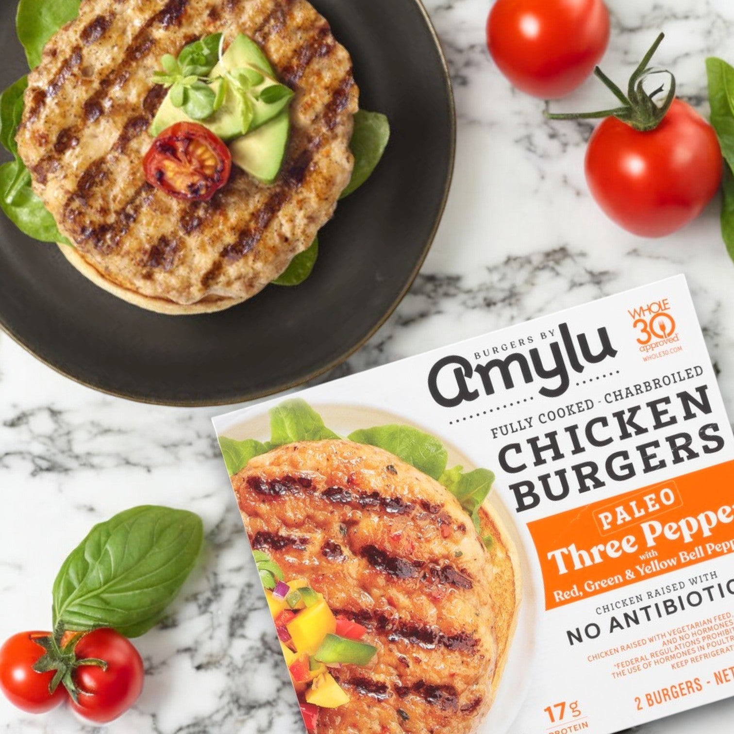 An 8 oz. package of Amylu Paleo Three Pepper Chicken Burgers sits on the table, showcasing a charbroiled burger with ripe avocado and cherry tomato. Fresh tomatoes and basil surround the dish, creating a perfect gluten-free delight.