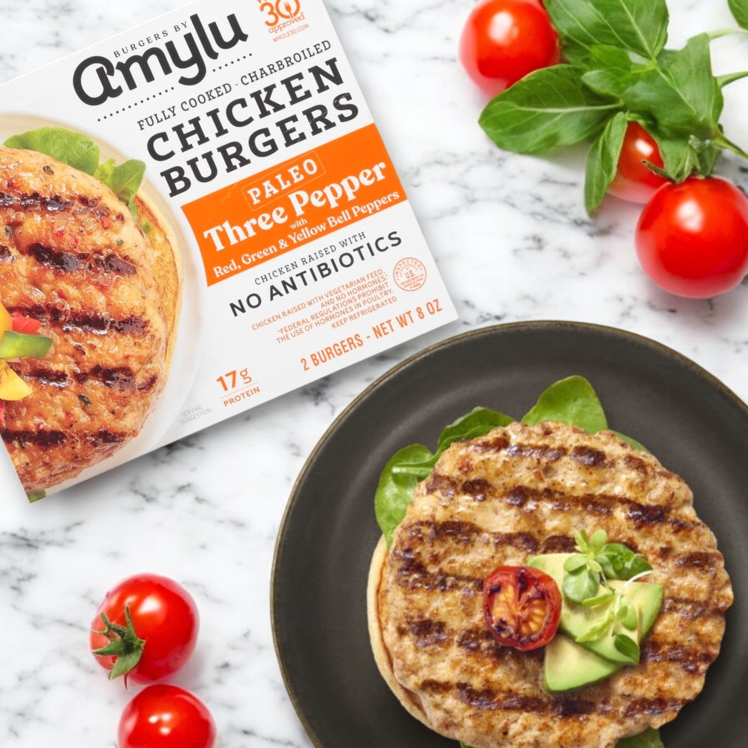 An Amylu Paleo Charbroiled Three Pepper Chicken Burger, grilled and topped with avocado and a cherry tomato slice, surrounded by whole cherry tomatoes and fresh herbs. Enjoy this Gluten-Free delight made from Antibiotic-Free Chicken.