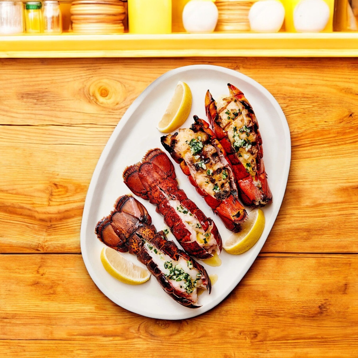 Member's Mark Warm Water Lobster Tails, sustainably sourced and garnished with herbs, are served with lemon wedges on a white plate atop a wooden table.