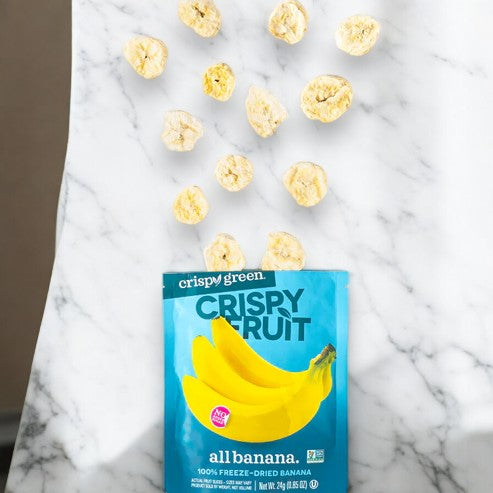 An open Crispy Green Crispy Fruit All Banana 0.85 oz packet lies on a marble surface with pieces spilling out. The blue packaging, by Crispy Fruit, showcases a banana illustration, promoting sustainable snacking.
