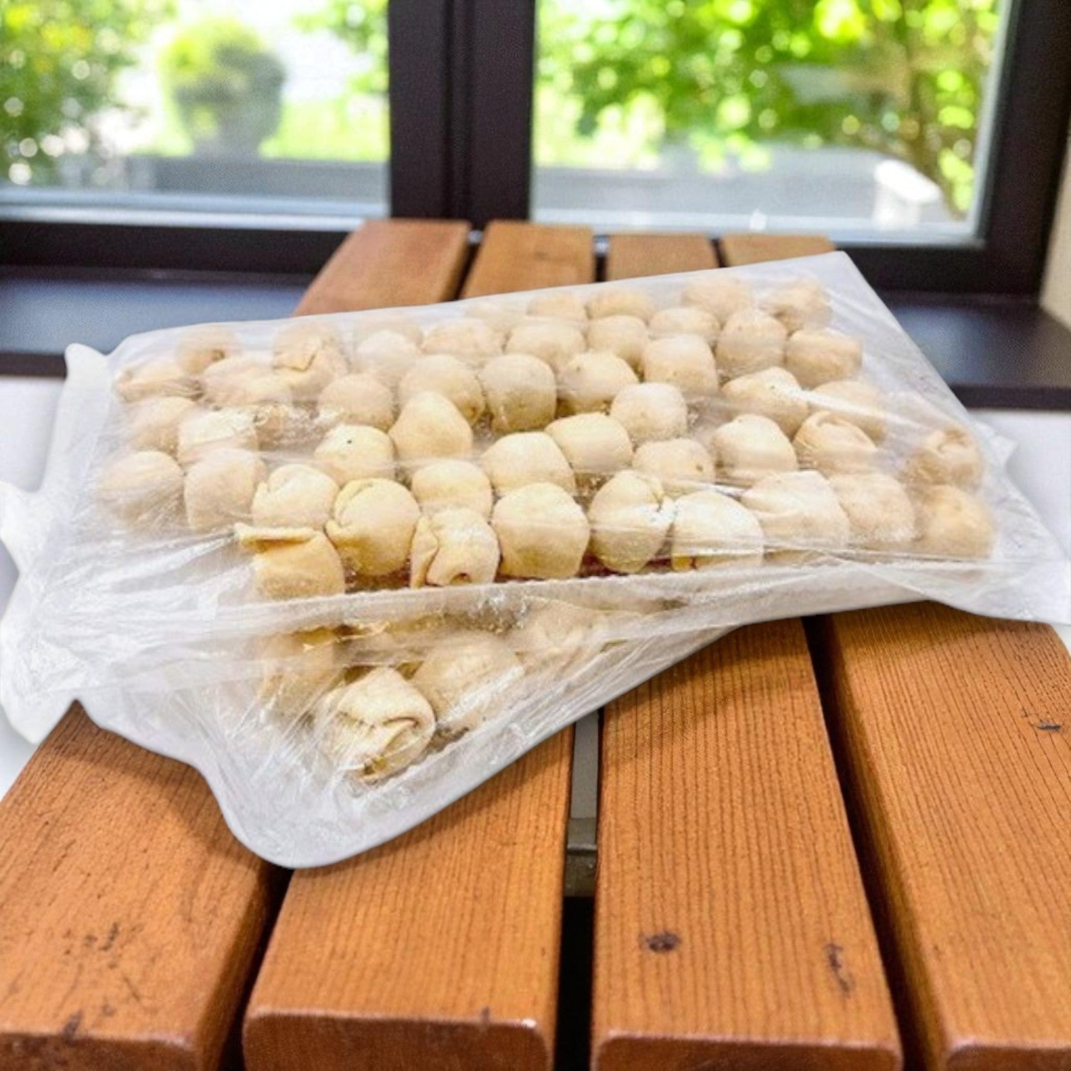 A vacuum-sealed pack of Van Lang Mini Beef Wellington (200 count, frozen appetizers) sits on a wooden bench near a window with greenery visible outside.