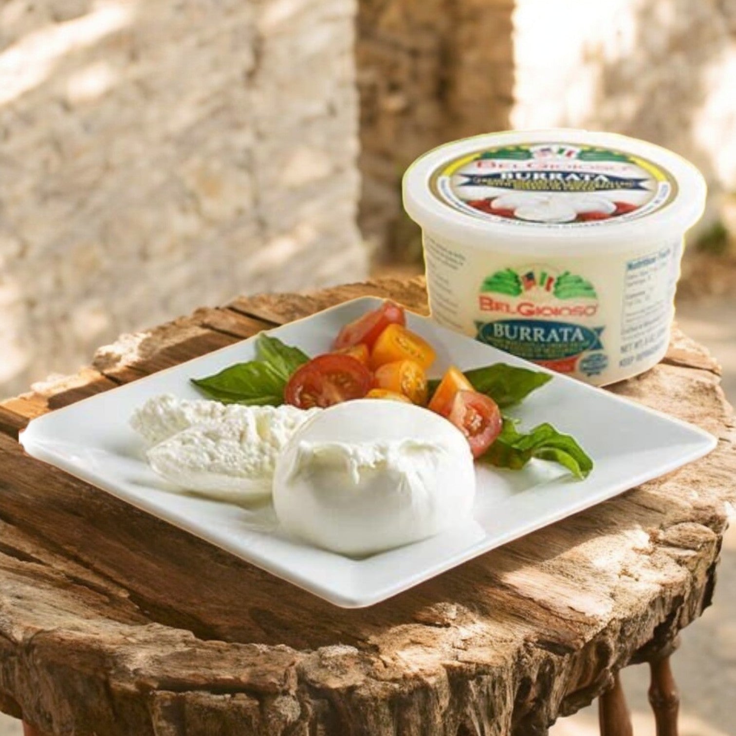 A rustic wooden table holds a plate with burrata cheese, tomato slices, and basil. A container of BelGioioso Fresh Mozzarella Burrata – 4 Ounce – 6/1 LB suggests an authentic Italian cheese delight in every bite.