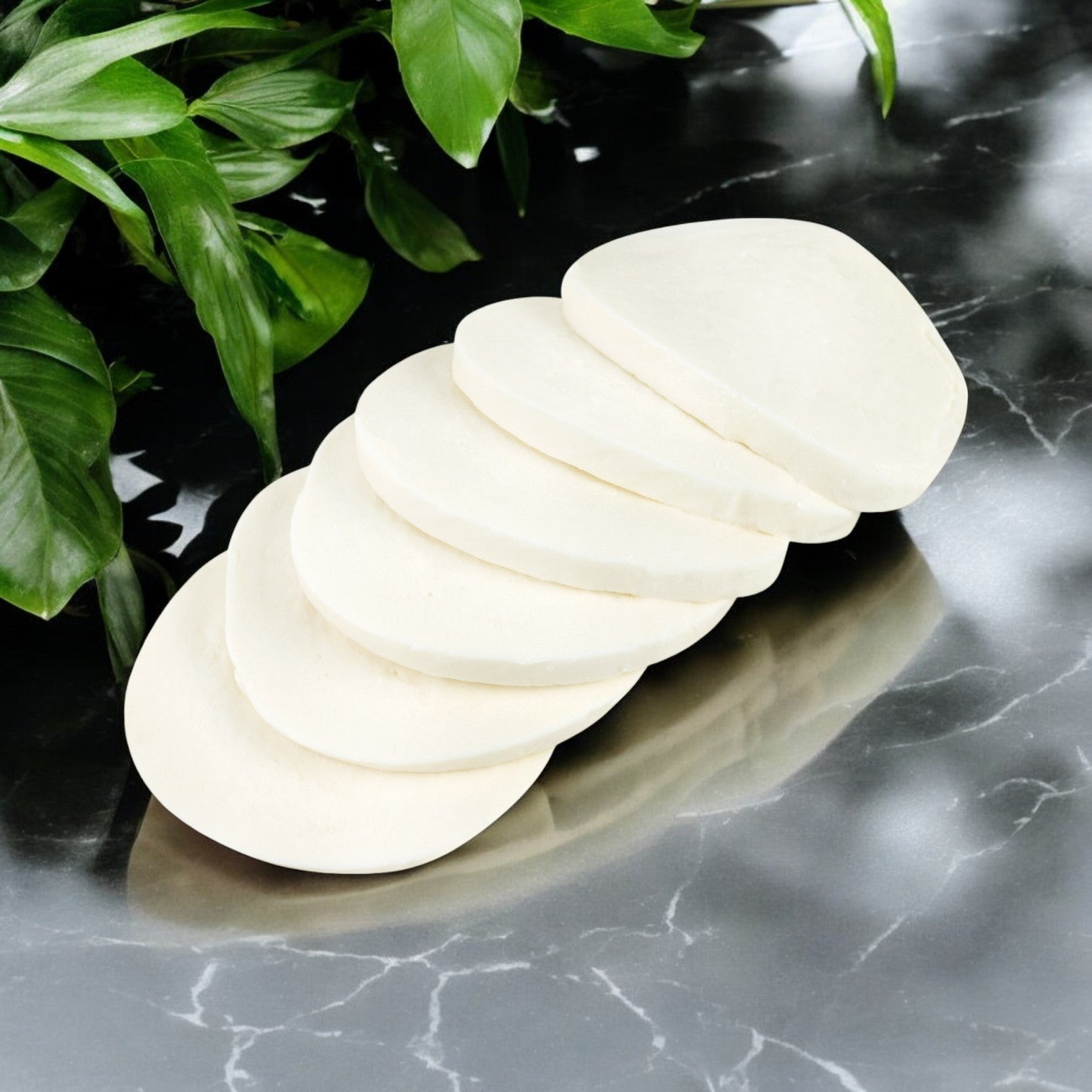 A cascading stack of six smooth, oval, white soap bars on dark marble mirrors the elegance of Belgioioso Fresh Mozzarella Cheese Thin Sliced. Green leaves create a lush backdrop.