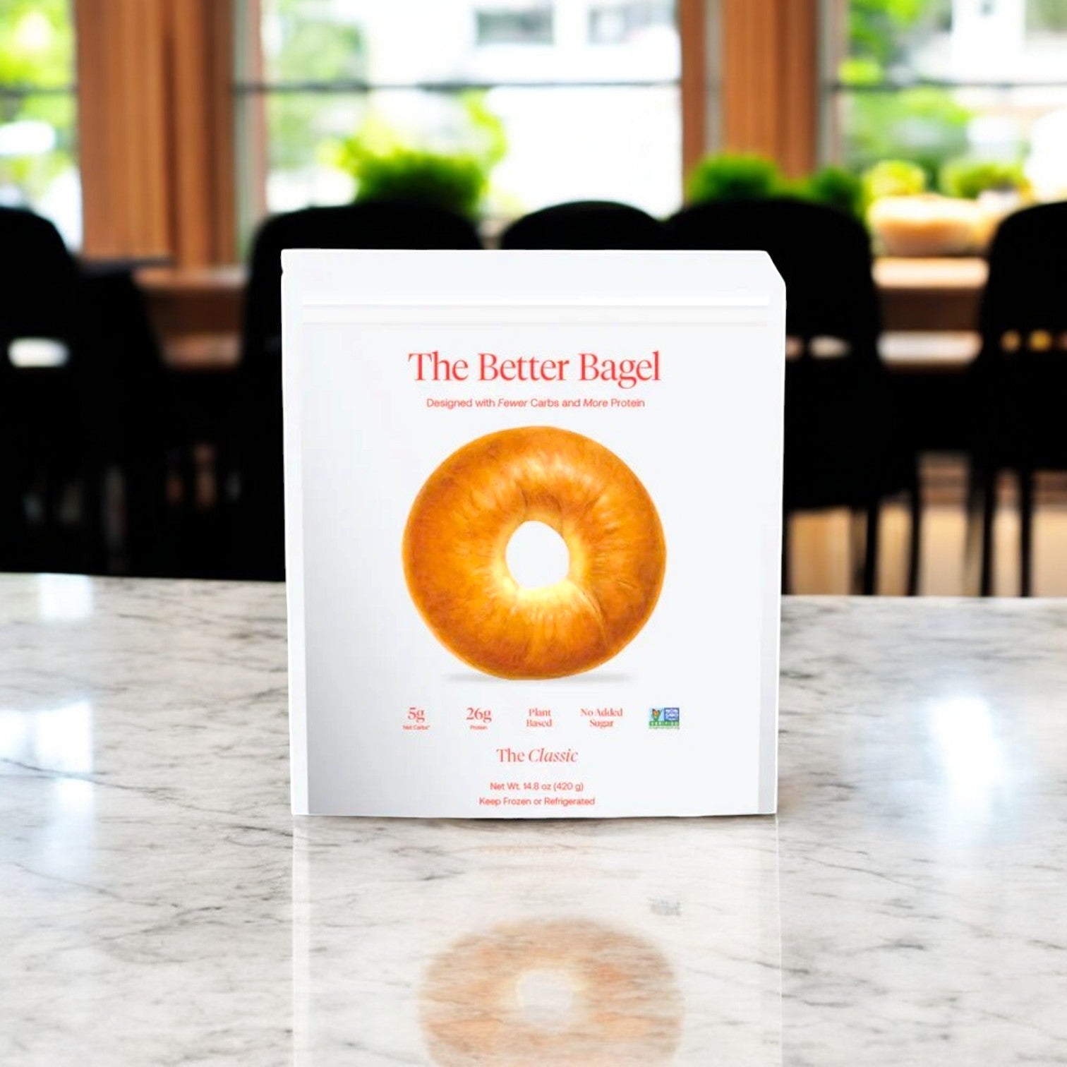 A box of "BetterBrand - Better Brand Variety Pack" rests on a marble counter, with blurred chairs and windows behind it. The packaging emphasizes lower carbs and higher protein.