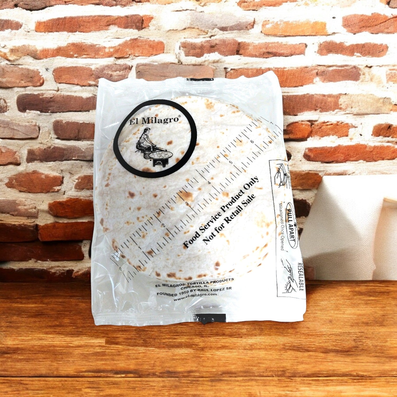 An El Milagro Flour Tortillas 10-inch pack graces a wooden surface, enhancing the scene with authentic flavor against a brick wall backdrop.