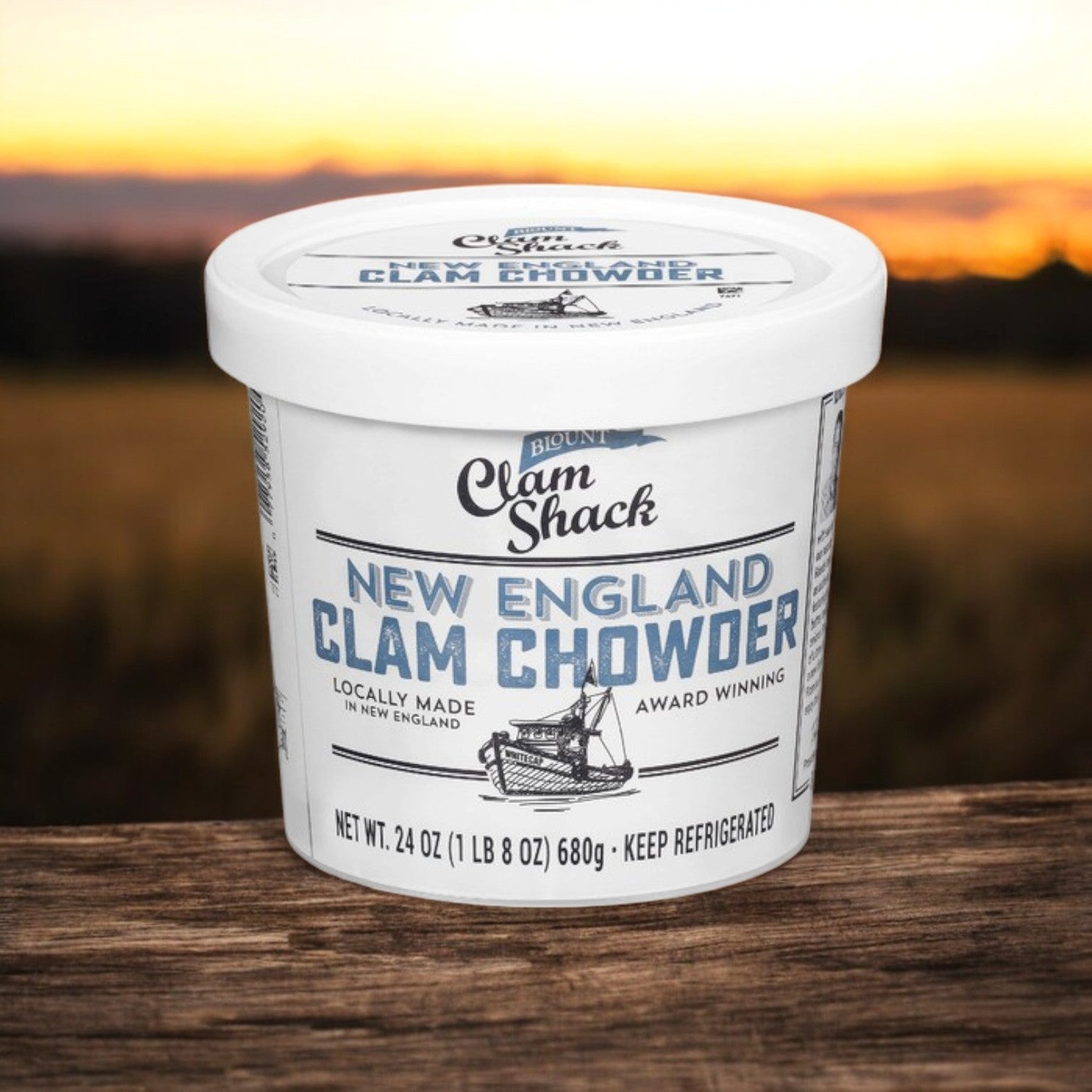 A white container of Blount Clam Shack Gluten Free New England Clam Chowder Soup - 24 oz., from Blount Fine Foods, made with premium ingredients, sits on a wooden surface with a sunset in the background.