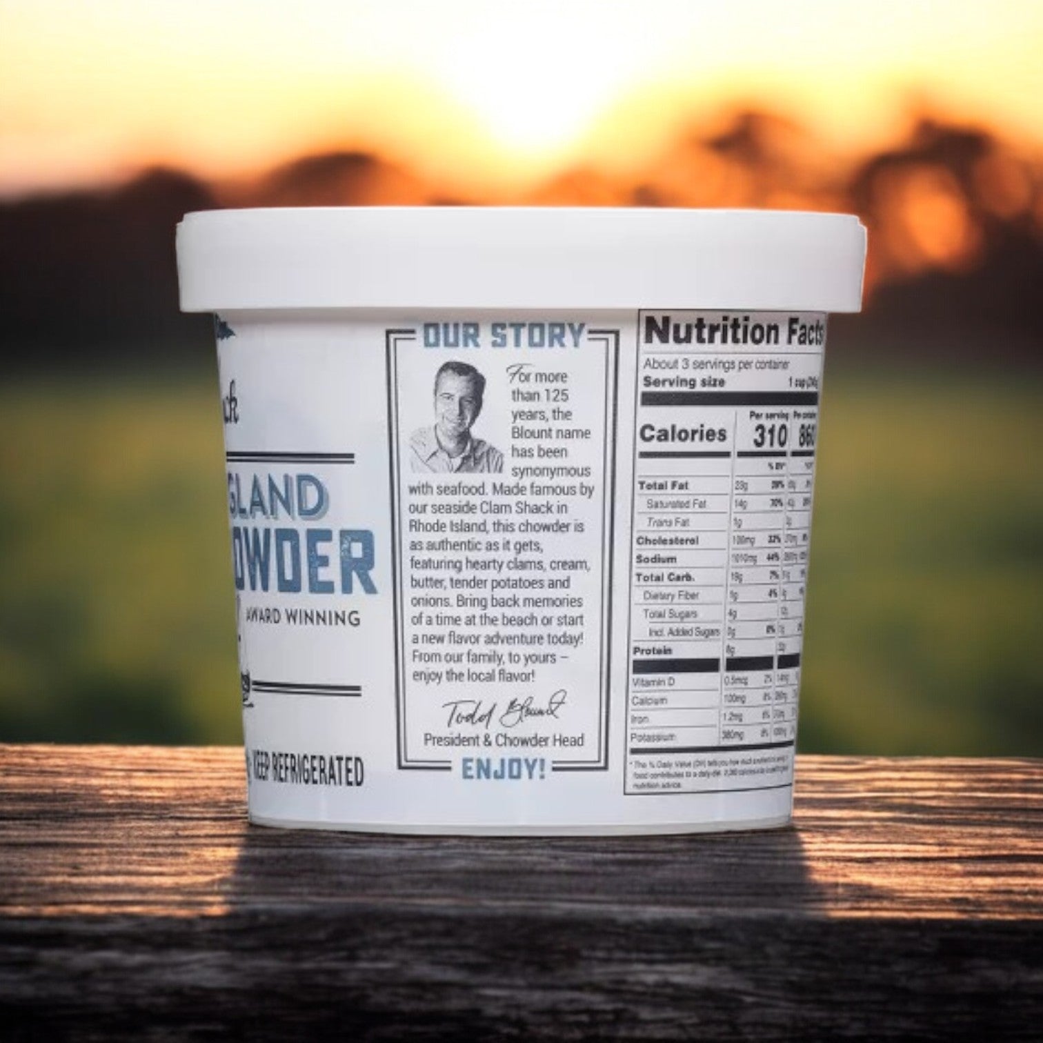 A 24 oz. container of Blount Clam Shack Gluten-Free New England Clam Chowder Soup from Blount Fine Foods rests on a wooden surface, featuring nutrition facts and brand story on the label, all with a serene sunset as the backdrop.