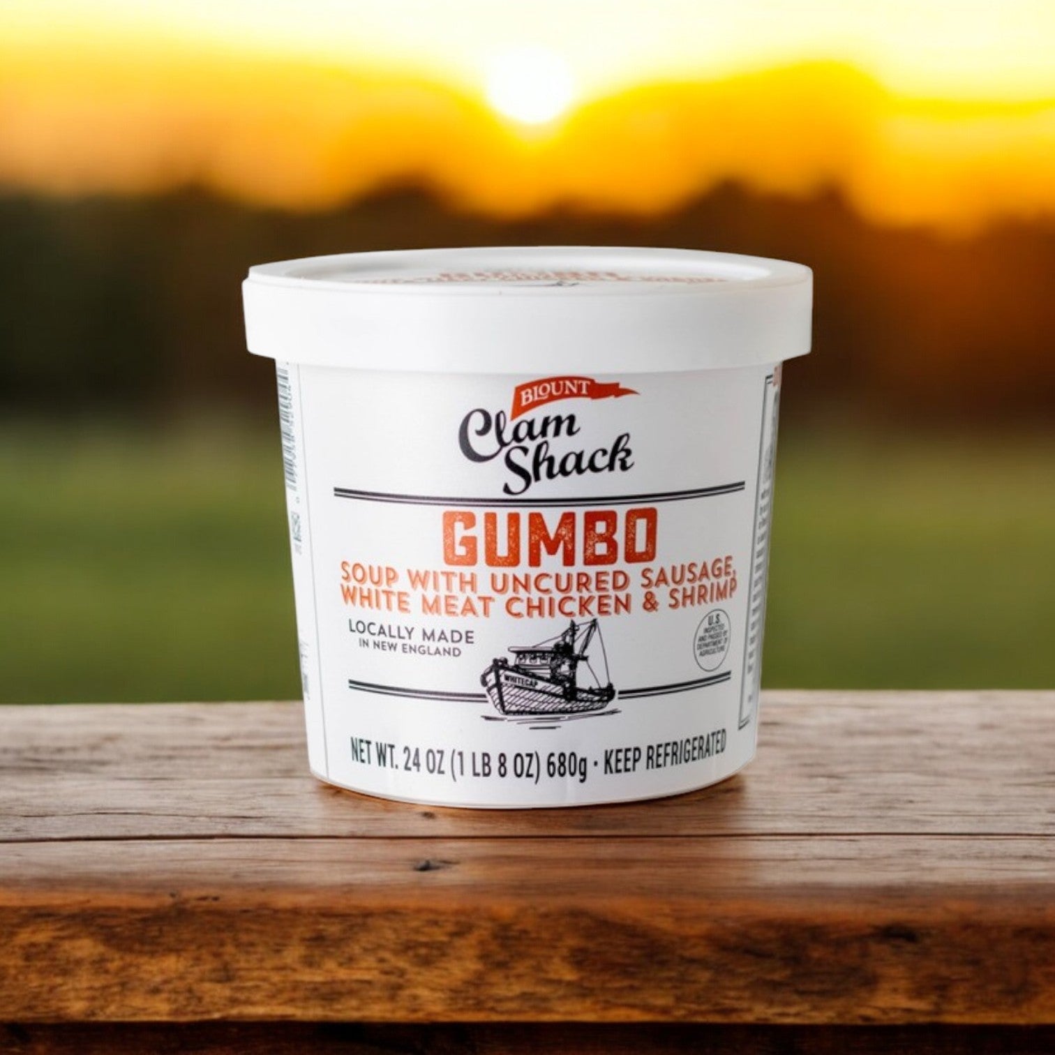 A 24 oz. container of Blount Fine Foods' Clam Shack Gumbo with uncured sausage, white meat chicken, and shrimp sits on a wooden surface against a brilliant sunset.