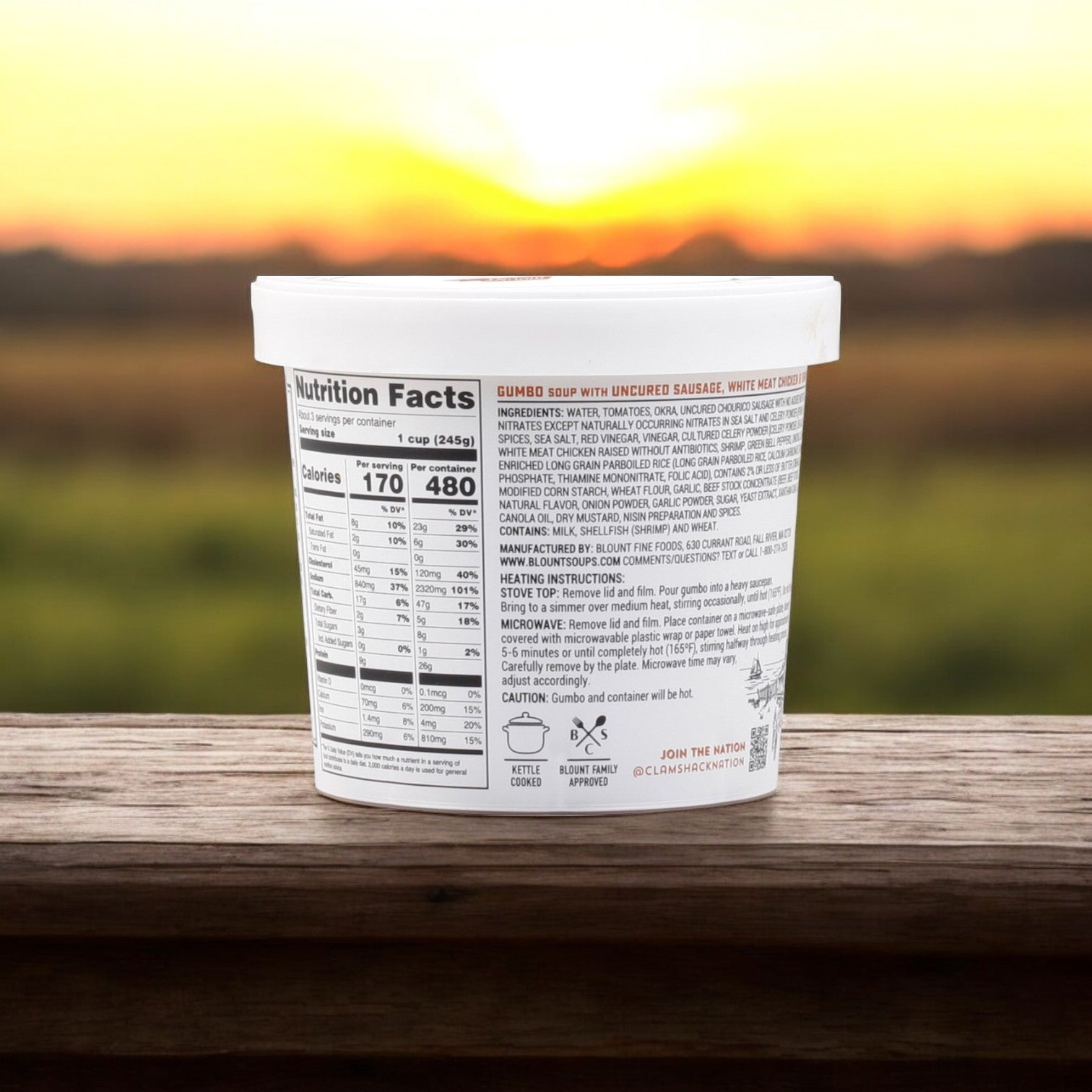 A 24 oz. container of Blount Clam Shack Gumbo with Uncured Sausage, White Meat Chicken & Shrimp Soup by Blount Fine Foods, featuring Creole spices in its ingredients and nutrition facts on the label, rests on a wooden surface against a sunset backdrop.