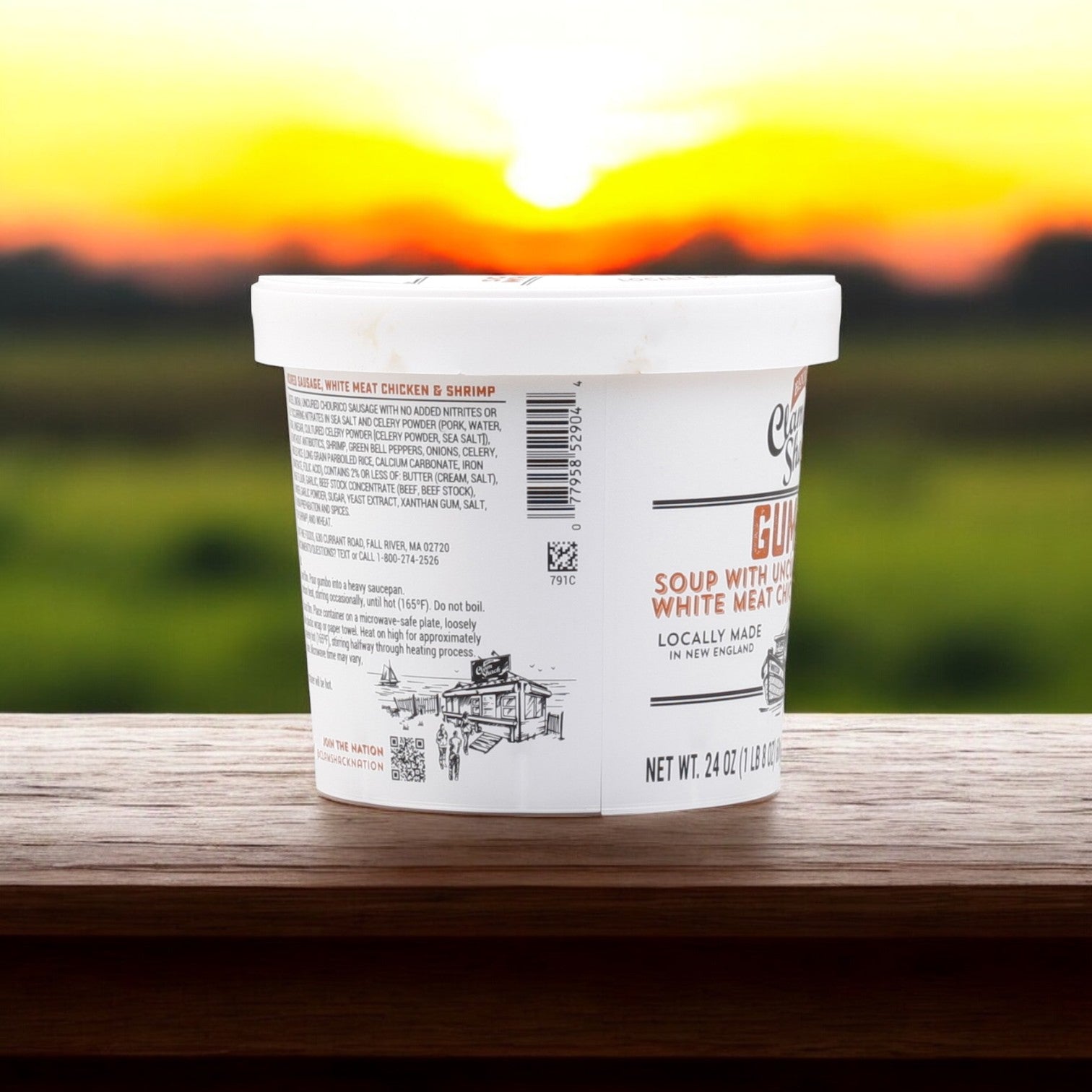A container of Blount Fine Foods' Clam Shack Gumbo with uncured sausage, white meat chicken, and shrimp soup, 24 oz., rests on a wooden surface against a sunset and grassy field.